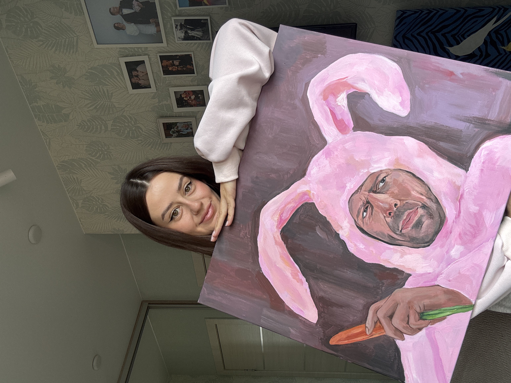 A woman holds an oil painting in front of her. The painting depicts a man in a pink bunny costume holding a carrot. The woman has dark hair and fair skin, and she is wearing a light pink sweatshirt. She is smiling and looking at the camera. The background includes a wall with a floral wallpaper and several framed photos. The location is Krasnoyarsk, Russia.