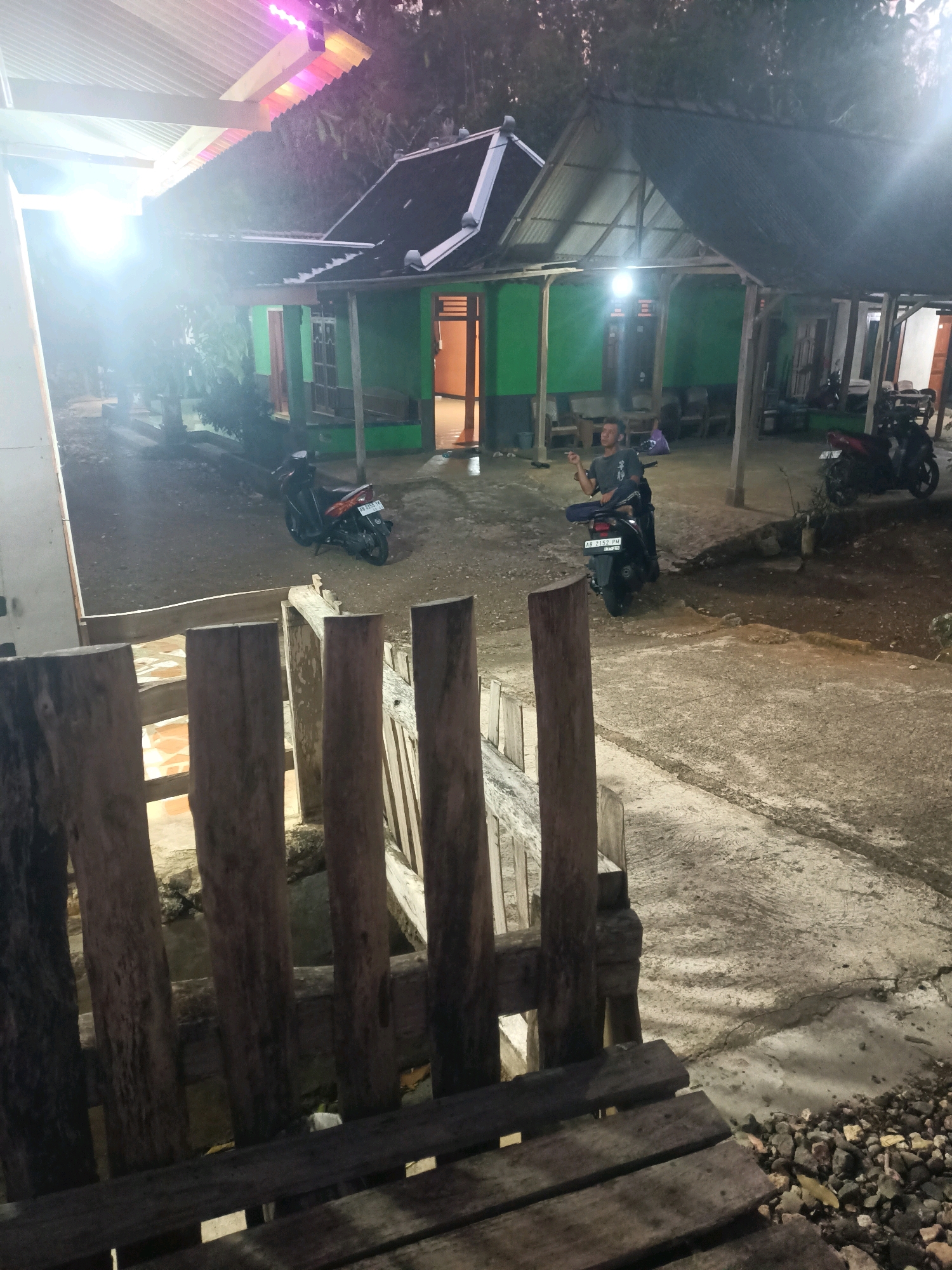 The image depicts an outdoor scene at dusk or night in Saptosari, Indonesia, with a rural or semi-rural residential setting. In the foreground, a rustic wooden bench and railing are visible, suggesting a raised vantage point overlooking a pathway. The pathway itself is made of concrete and gravel, with visible cracks and rough texture.
Behind the pathway, two motorcycles are parked. One is a dark-colored scooter with a license plate that reads "AB 2157 PM". The second motorcycle, partially visible, appears to be red and white.
A man is seated on the dark scooter, wearing a dark t-shirt and appears to be holding a cigarette or small object in his right hand. He is looking towards the right of the frame.
The background features a single-story green building with a dark corrugated metal roof, supported by wooden beams. The building has open doorways revealing interior spaces. One doorway shows what appears to be an orange-colored wall. Lights illuminate the area, including a bright light source emanating from the left side of the frame, creating a lens flare. Colored lights, possibly part of a decorative lighting system, are visible on the edge of a roof structure in the upper left corner. Trees and foliage surround the building, contributing to a natural environment. The overall atmosphere is dim, suggesting it is after sunset.