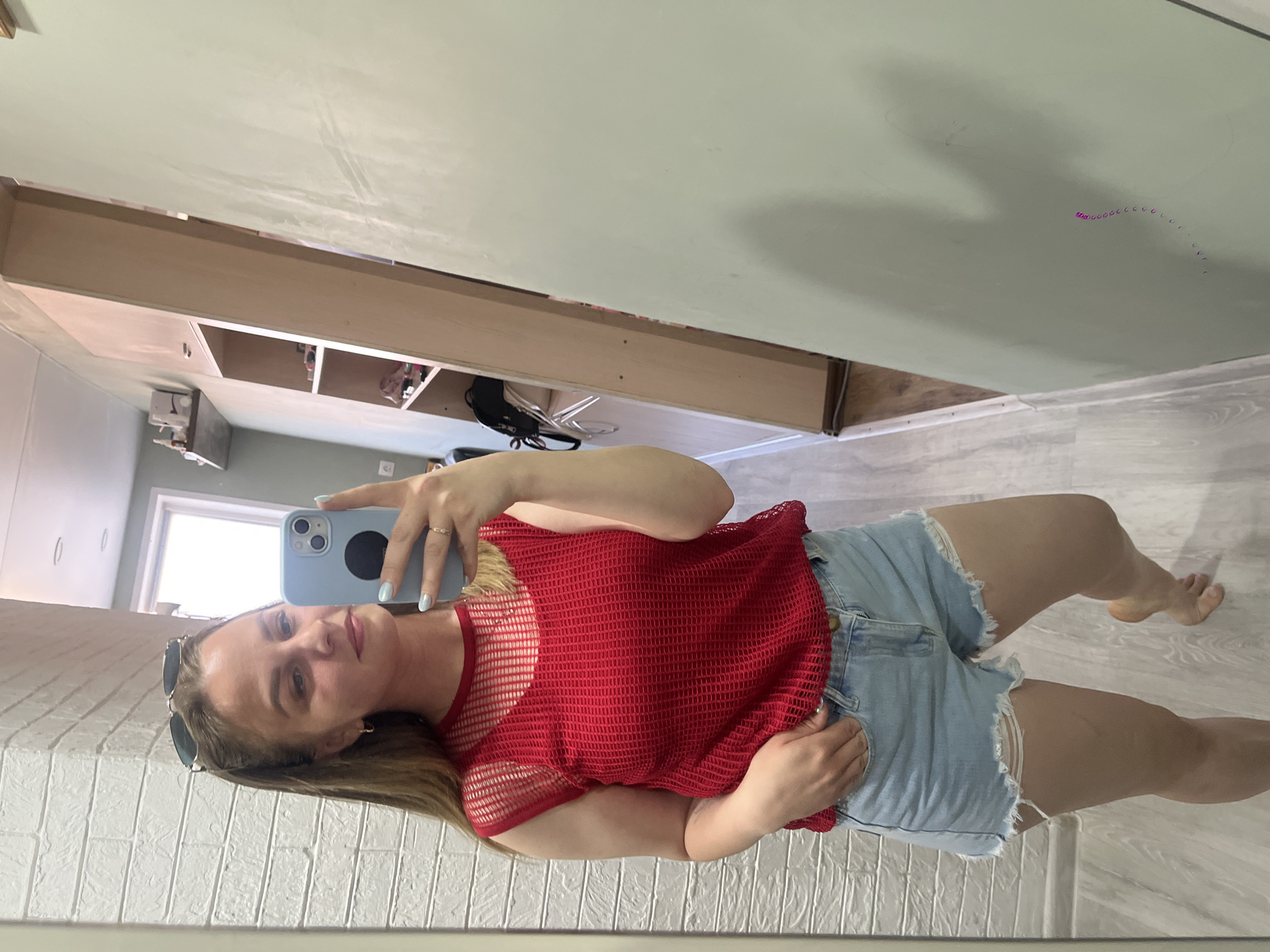 A young woman stands in front of a mirror taking a selfie with a light blue smartphone. She wears a red, net-like, short-sleeved shirt and distressed denim shorts. The environment appears to be indoors, with a white brick-like wall behind the mirror, and a shelf unit and green wall visible in the mirror's reflection. The location is Izhevsk, Russia.