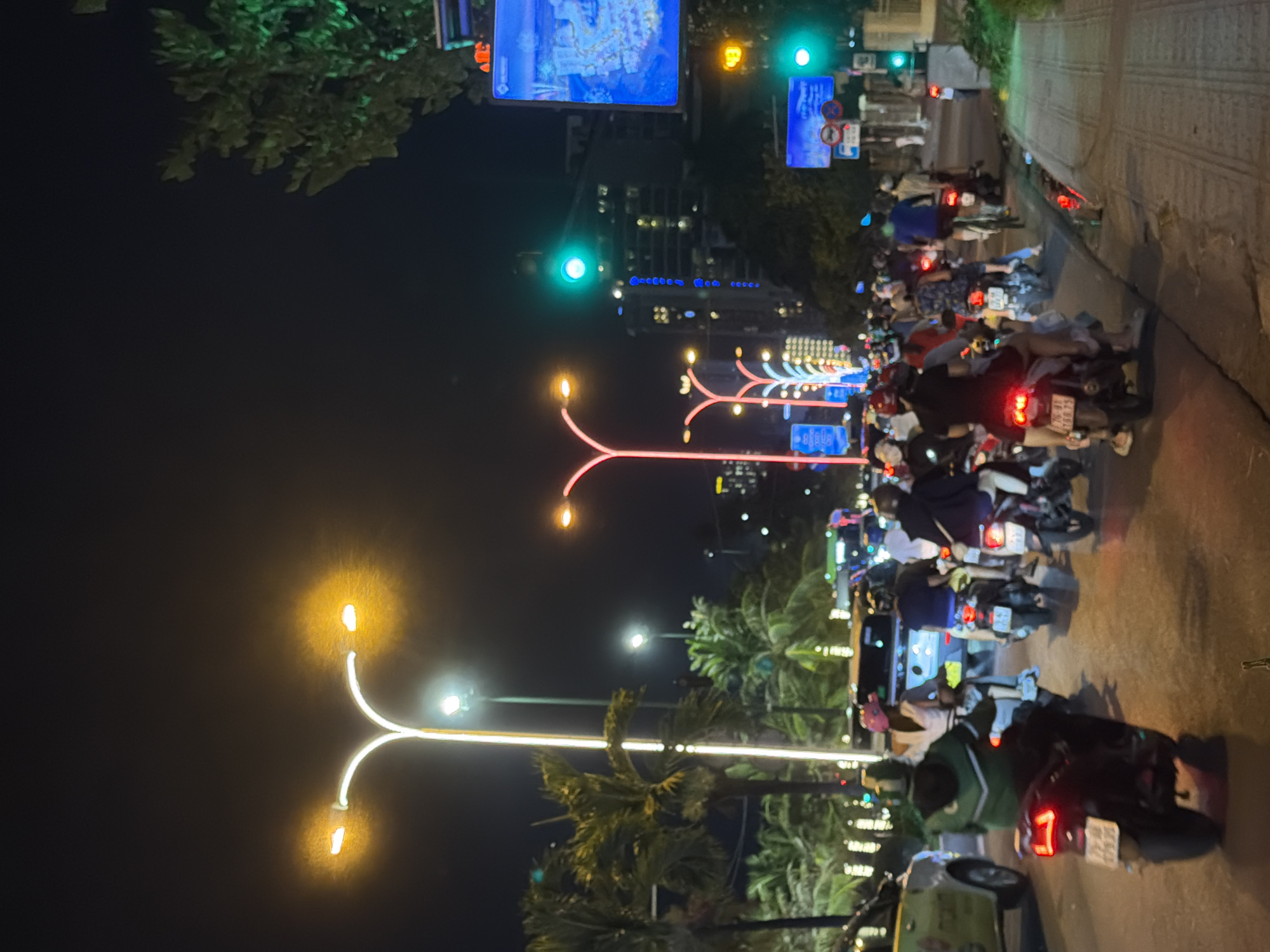 This image captures a lively night scene in Nha Trang, Vietnam, likely on a bustling street. The photograph is oriented vertically. The dominant subjects are numerous people riding on motorbikes, forming a significant traffic flow. They are dressed in casual attire, some wearing helmets.
The setting appears to be an urban environment at night, illuminated by artificial lights. Various light sources are visible, including streetlights, decorative lights forming arches over the road, traffic signals (green and yellow lights are discernable), and the headlights and taillights of the motorbikes and other vehicles. Palm trees are scattered along the roadside, suggesting a tropical or sub-tropical climate. The road surface is paved, and a pedestrian walkway is visible to the right, with a patterned tile design.
The activity depicted is the movement of traffic, primarily motorbikes, suggesting a busy evening. The presence of many vehicles and people implies a vibrant, active atmosphere. The time of day is clearly night, indicated by the darkness and the reliance on artificial lighting.
Notable details include the bright, colorful decorative lighting and the density of motorbikes, which are a common mode of transportation in Vietnam. There are no explicit emotional cues, but the bustling activity suggests a dynamic and energetic scene.
Visible text includes license plates on some of the motorbikes, which appear to be in Vietnamese script and numerals. Additionally, there are signs with text and symbols, including what seem to be traffic signs, and possibly advertisements or informational displays on larger screens mounted on poles. One sign clearly displays the number "49," potentially indicating a route or building number. The overall impression is of a vibrant, energetic city at night.