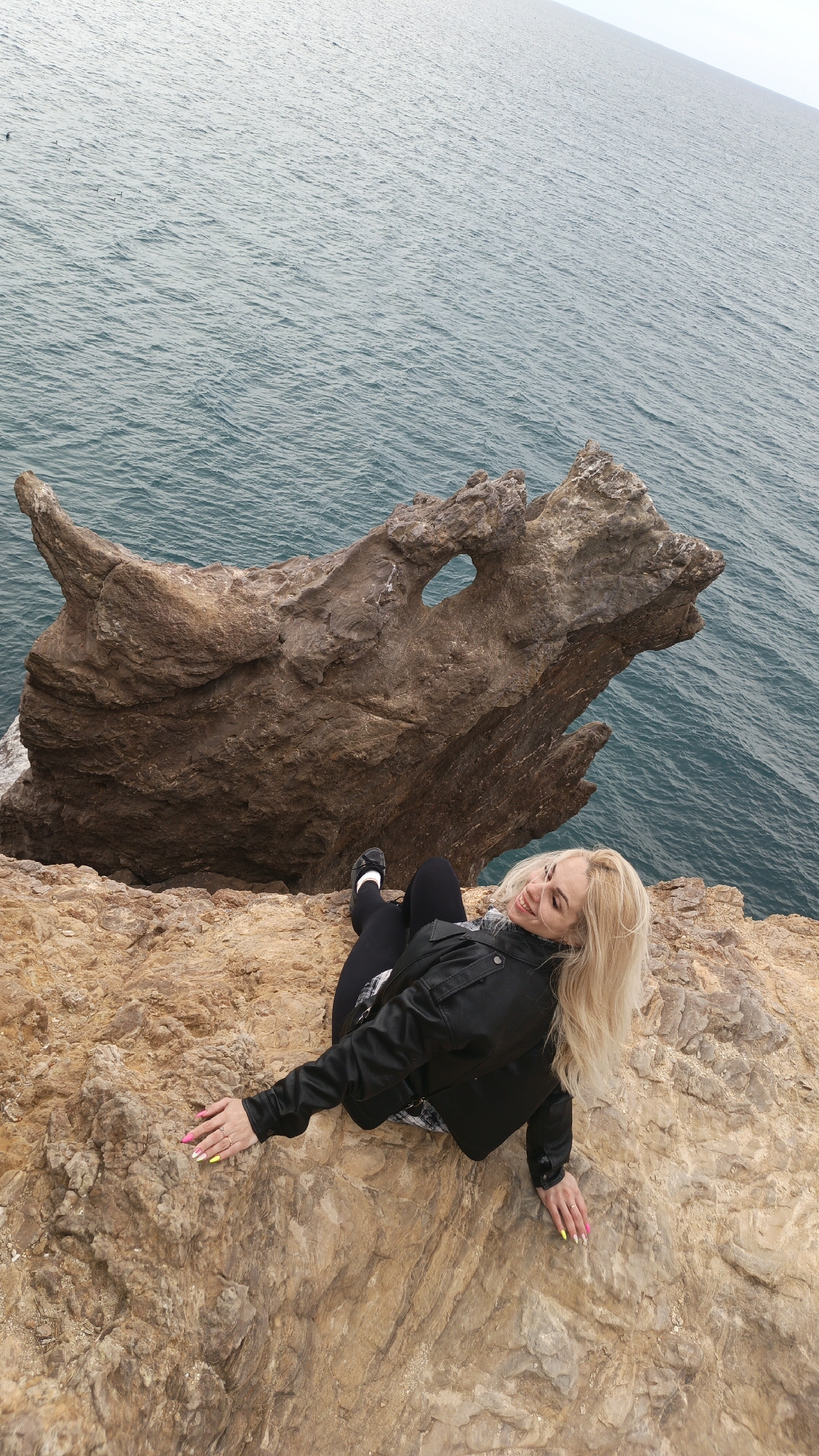 A high-angle shot captures a blonde woman lying on a rocky cliff edge overlooking the sea. The setting is a rugged coastline, likely in Балаклавский округ, Ukraine. The woman is positioned in the lower right and center of the frame, smiling and looking up towards the camera. She is dressed in a black leather jacket, a plaid or checkered shirt underneath, black leggings, and black shoes with white socks. Her long, blonde hair cascades around her head. Her left hand, adorned with rings and brightly colored pink and yellow nail polish, rests on the textured brown rock of the cliff.
To her left, a large, distinctive brown rock formation protrudes over the water, resembling a dragon's head or a similar mythical creature, complete with a prominent hole that suggests an eye or nostril. The cliffside is composed of rough, earthy-toned rocks. Beyond the cliffs, the expansive, calm waters of the sea stretch out to the horizon under a soft, overcast sky, giving the water a muted grey-blue hue with subtle ripples. The overall impression is one of natural beauty and serene enjoyment. There is no visible text in the image.