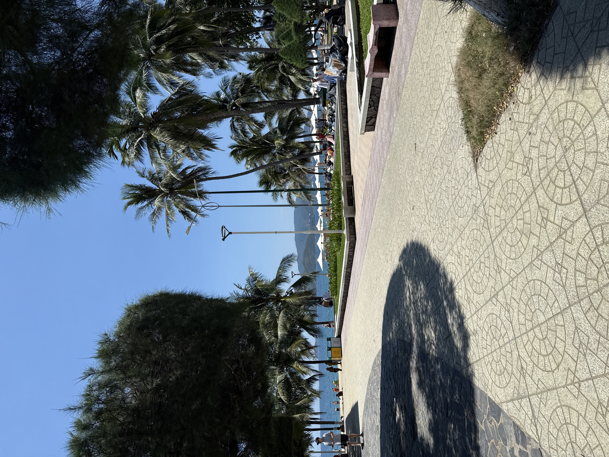 The image depicts a beachside promenade in Nha Trang Ward, Vietnam. Palm trees line the beach, and the blue ocean is visible in the background with distant mountains. A paved walkway with a geometric tile pattern runs alongside the beach. Benches are placed along the walkway, near patches of green grass. People can be seen walking and relaxing near the beach, many sitting under beach umbrellas. A tall street lamp stands along the path. A large shadow cast by a tree dominates the right side of the paved walkway.
