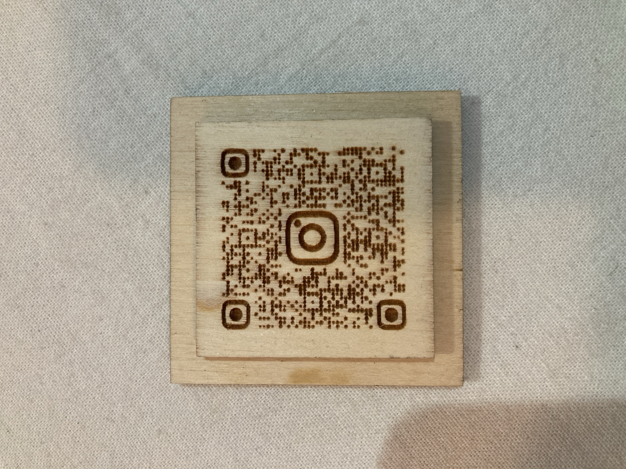 A wooden QR code with an Instagram logo in the center is placed on a white textured surface. The QR code is etched into the wood, and the Instagram logo is also visible within the code. The wooden QR code is slightly raised from the surface it rests on. The background is a neutral white fabric with a subtle texture.