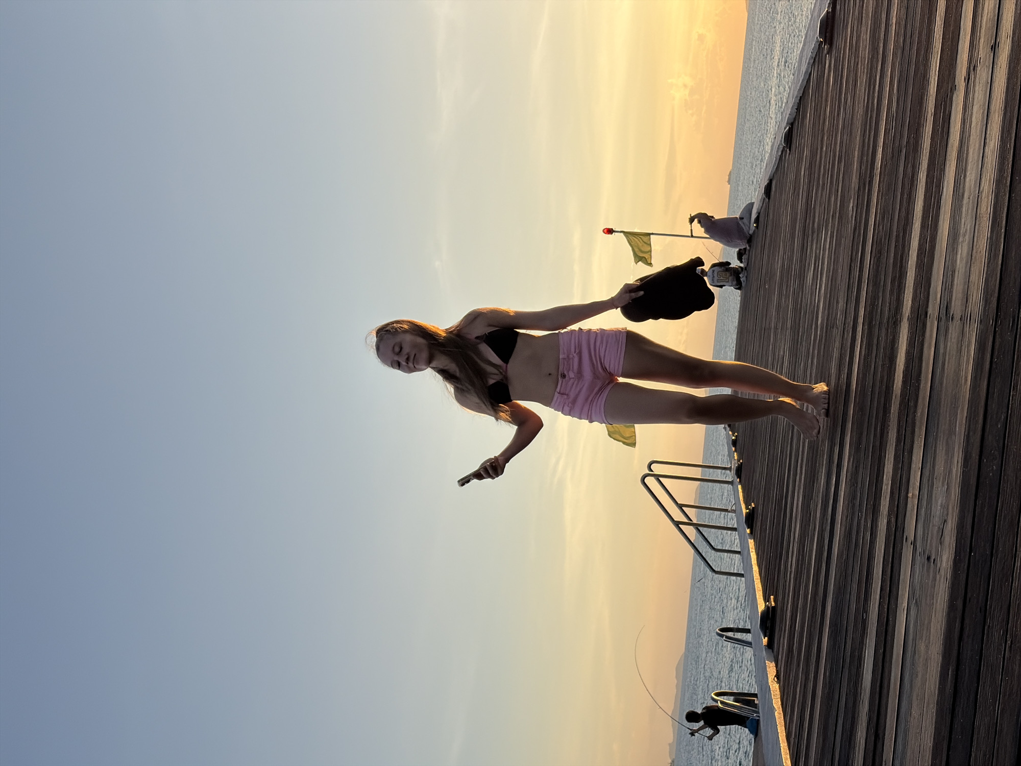 A young woman stands on a wooden pier in Baan Taling Ngam, Thailand, at sunset. She is wearing a black bikini top and pink shorts, and is holding a black hat in her left hand while looking at her cell phone in her right hand. The sky is a gradient of blue to orange. In the background, several people are visible on the pier, including a person fishing and two people sitting. The ocean stretches out to the horizon, with islands visible in the distance. The wooden planks of the pier are illuminated by the setting sun.