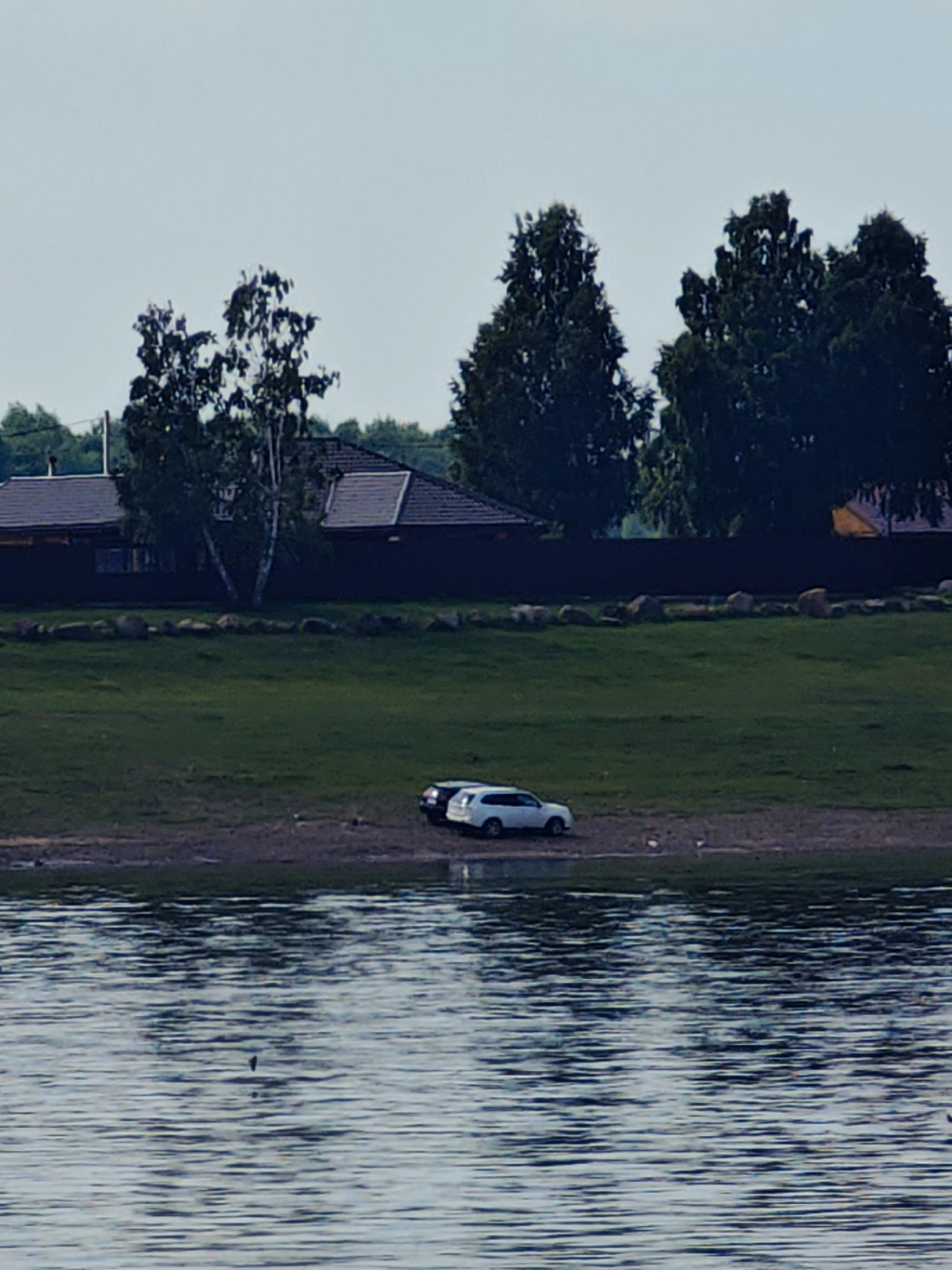 The image captures a serene waterscape with a shoreline dotted with two parked cars. The larger, white car, possibly a SUV, is positioned on the right side of the frame, while a dark-colored car is situated to its left. The cars are parked on a strip of dry land adjacent to a grassy embankment. This embankment leads up to a residential area, characterized by dark-paneled houses with prominent brown roofs. A line of large, rough-hewn stones marks the transition between the embankment and the residential area. The background is filled with dense foliage, dominated by tall, dark green trees, suggesting a rural or suburban setting. The sky above is a pale, cloudless blue. The foreground of the image is the surface of a body of water, showing subtle ripples and reflections, giving the impression of a calm day. While the specific location is identified as Erkalovo, Russia, the image itself provides no further context about human activity beyond the parked vehicles.