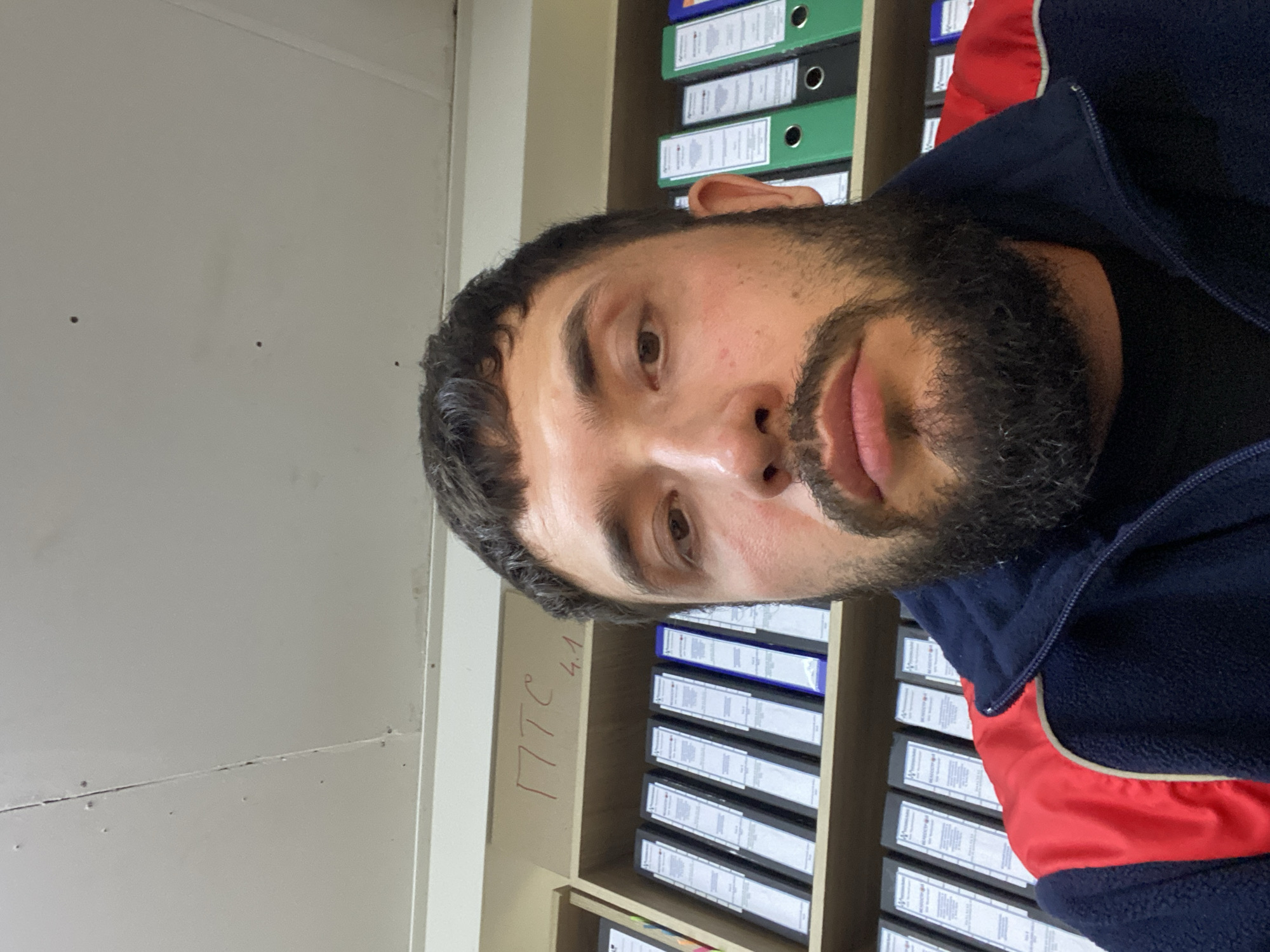 The image is a selfie taken by a man with dark hair and a beard, wearing a dark blue jacket with red accents. He is looking directly at the camera with a neutral expression. Behind him, a shelf filled with binders is visible. The shelf has some writing in red marker that appears to read "ПТС 4.1". The environment appears to be an office or storage area. The ceiling above is white and shows some signs of wear. The man is positioned in the foreground, with the shelves and binders slightly out of focus in the background. The lighting is bright, suggesting indoor illumination. The image's context places it in Luzhitsy, Russia.