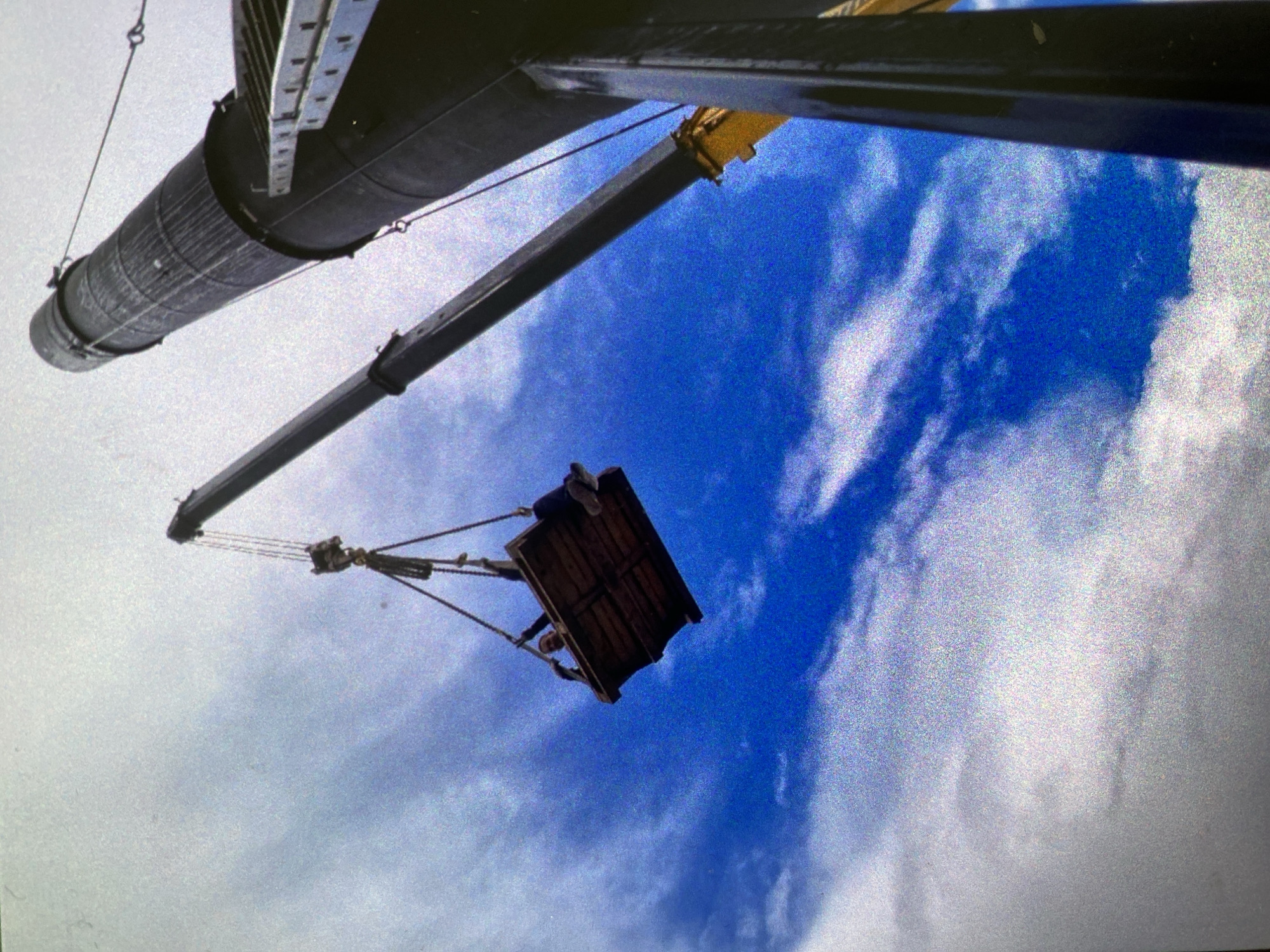 The image presents an upward-looking view of a large crane in operation against a bright blue sky filled with scattered white clouds. Dominating the upper left portion is a substantial, dark, cylindrical structure, possibly a component of a larger crane or an industrial stack, detailed with horizontal bands and a ladder-like structure running along its top edge.
Extending from the upper right, a dark crane arm with a visible yellow joint descends into the frame. Suspended by cables from this crane arm is a dark wooden platform or cage. On this platform, at least one individual is visible, appearing to be sitting or crouching, with their legs and feet clearly seen.
The scene captures the activity of a person being hoisted or lowered by a crane on a work platform, likely engaged in construction, maintenance, or inspection work on a tall structure. The time of day is clearly daytime, characterized by the bright sky and fair weather. The image has a noticeable grainy quality, suggesting it may be a photo of a screen or a scan of an older print. No text is visible within the image. The location context for this activity is Saint Petersburg, Russia.