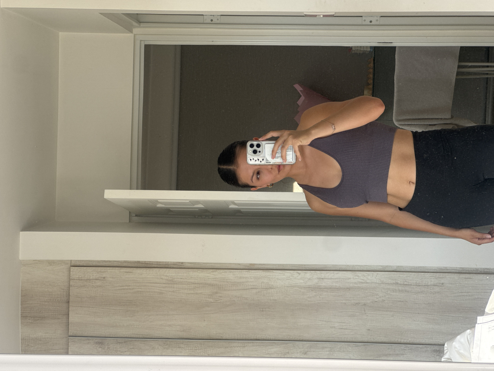 The image is a mirror selfie showing a young woman indoors, presented with a 90-degree clockwise rotation. The woman, who appears to be standing upright in the reflection, has dark hair pulled back and is wearing a dark purple or gray ribbed crop top and dark leggings. She accessorizes with small hoop earrings and a delicate silver bracelet on her left wrist. She is holding a smartphone, likely an iPhone given the camera configuration, encased in a clear cover adorned with small star stickers, as she takes the photo. Her expression is neutral as she looks towards the phone's camera.
The background, visible through the mirror's reflection, depicts an indoor setting. A light-colored wall and a white door frame are seen behind her, along with a dark floor or carpet. Further back, a gray towel or fabric is draped over an object. In the foreground of the reflection, a modern piece of furniture or cabinetry with a light, wood-grain texture is visible. The lighting is diffuse, suggesting an interior space without direct sunlight. There is no clearly legible text other than the decorative stars on the phone case.