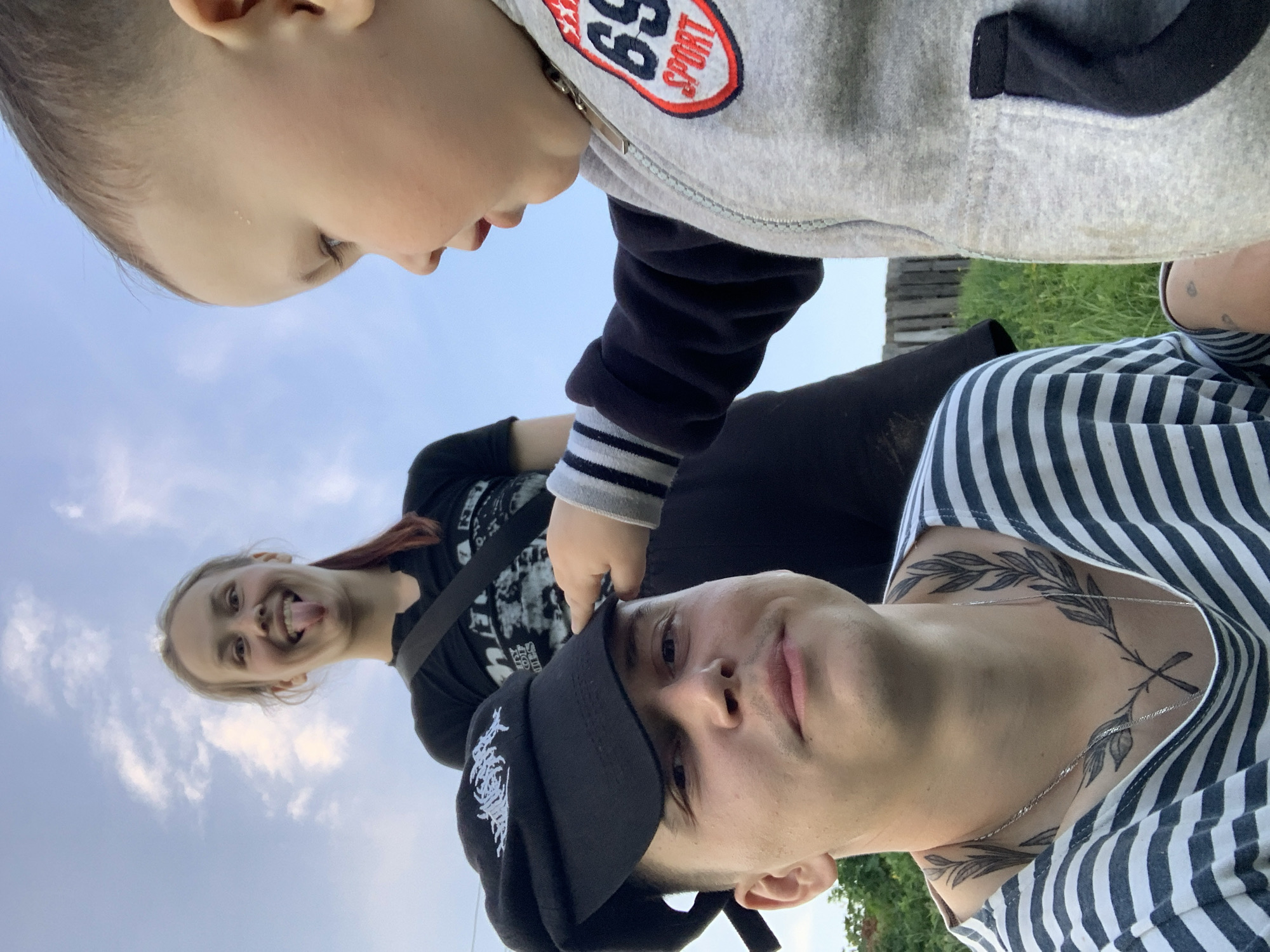 A low-angle shot captures a group of three individuals. The foreground features a man in a black and white striped shirt with a neck tattoo and a black cap with white embroidery. A child in a grey sweatshirt with a "69" emblem stands above him, looking down. In the background, a woman wearing a black t-shirt smiles with her tongue sticking out. They appear to be outdoors, possibly in Фоки, Russia, based on the prompt. The sky is visible in the background.