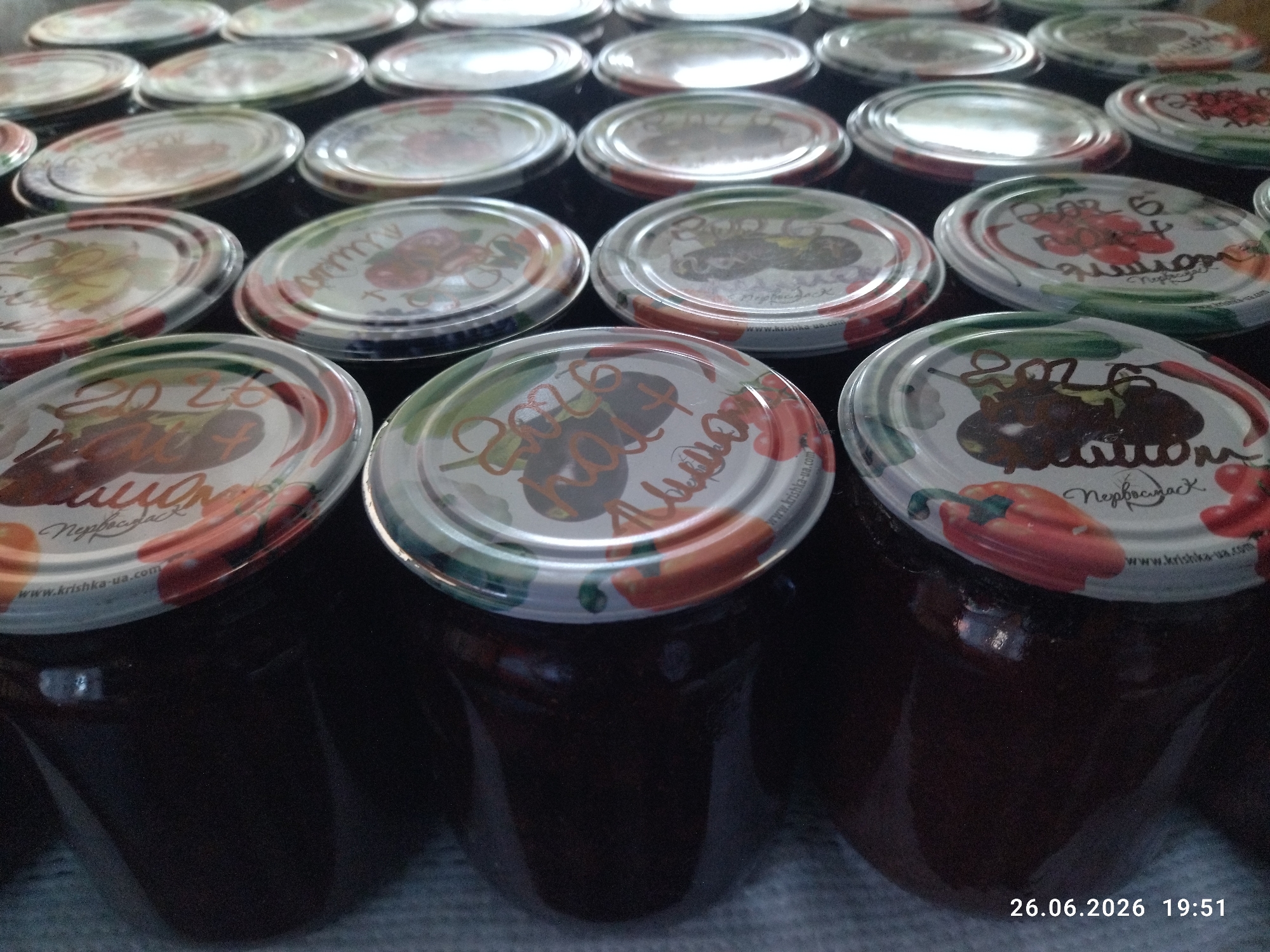 The image displays a close-up, overhead view of numerous jars of preserves, likely homemade, arranged in a grid. The jars have metal lids adorned with colorful illustrations of fruits and vegetables, and some also feature text. The preserves themselves are dark in color, appearing to be jam or jelly. The lids have a metallic sheen, reflecting the light source.
Several jars prominently feature the year "2026" written in an orange script, suggesting they were made in that year. One lid has the Cyrillic text "Нервбаст" and "www.krishka-ua.com", indicating a brand or origin. Another lid shows a drawing of peppers. The overall scene suggests a food preservation activity, likely taking place in a home or a small production setting.
The lighting is somewhat dim, with reflections on the jar lids indicating an indoor setting. The focus is sharpest on the jars in the foreground and middle ground, with those in the background becoming progressively blurred. The lower right corner of the image displays a timestamp: "26.06.2026 19:51," suggesting the photograph was taken on June 26, 2026, at 7:51 PM. The mention of Zavodske, Ukraine, provides a geographical context for this scene. There are no people visible in the image.