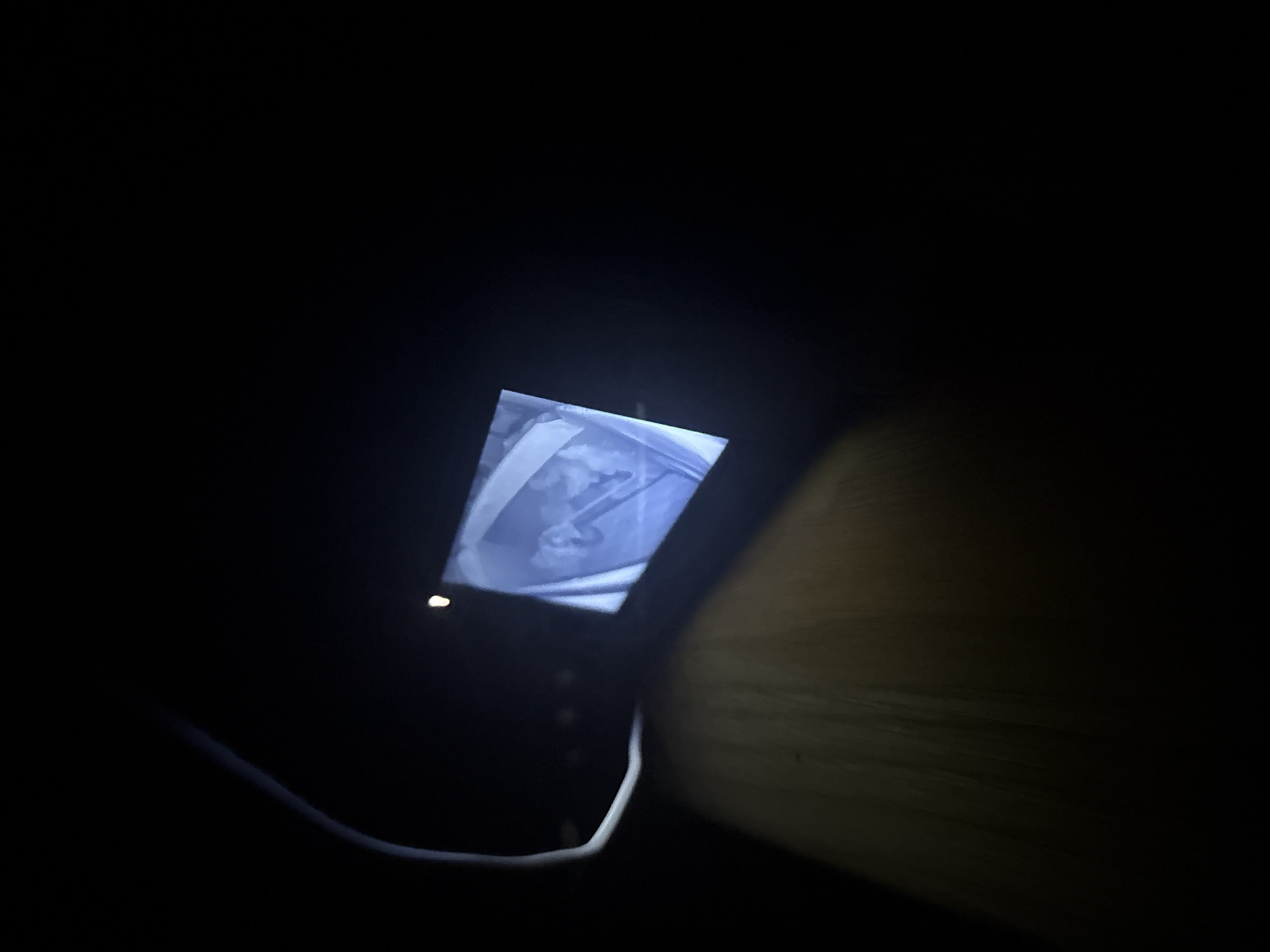 An electronic device with a glowing screen rests in a very dark environment. A cable is connected to the device. A wooden surface is visible nearby. The screen displays an image that appears to be a grayscale medical image. The exact location is unknown.