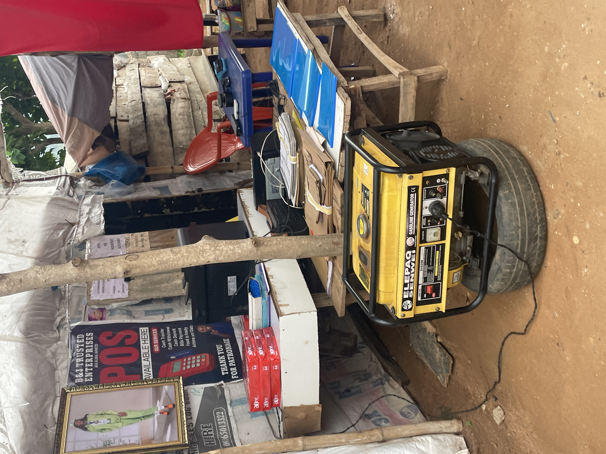 The image depicts an outdoor stall or shop in Lokoja, Nigeria, likely a small business operating in an informal setting. In the foreground, a yellow Elepaq Senwei gasoline generator sits on a spare tire, with a black electrical cord extending to the left. To the left of the generator is a wooden table with stacks of red A4 paper and some notebooks. Behind this table, a larger wooden structure holds more papers and blue plastic folders. Further back, a blue table holds a red plastic chair, a small black electronic device, and some stationery items.
The stall itself appears to be constructed with various materials, including tarpaulin roofing and wooden supports. A prominent poster on the wall behind the generator advertises "B&J TRUSTED ENTERPRISES" offering "POS AVAILABLE HERE" and listing services such as cash transfer, cash withdrawal, and bill payment. A framed picture of a person in a green tracksuit is displayed next to the POS poster. Other signs and posters are visible, some displaying contact information. The ground is a reddish-brown dirt. In the background, stacked wooden planks and a red cloth hanging down suggest further storage or partition. The overall scene conveys a busy, functional, and somewhat makeshift business environment characteristic of market stalls in Lokoja. There are no visible people in the image.