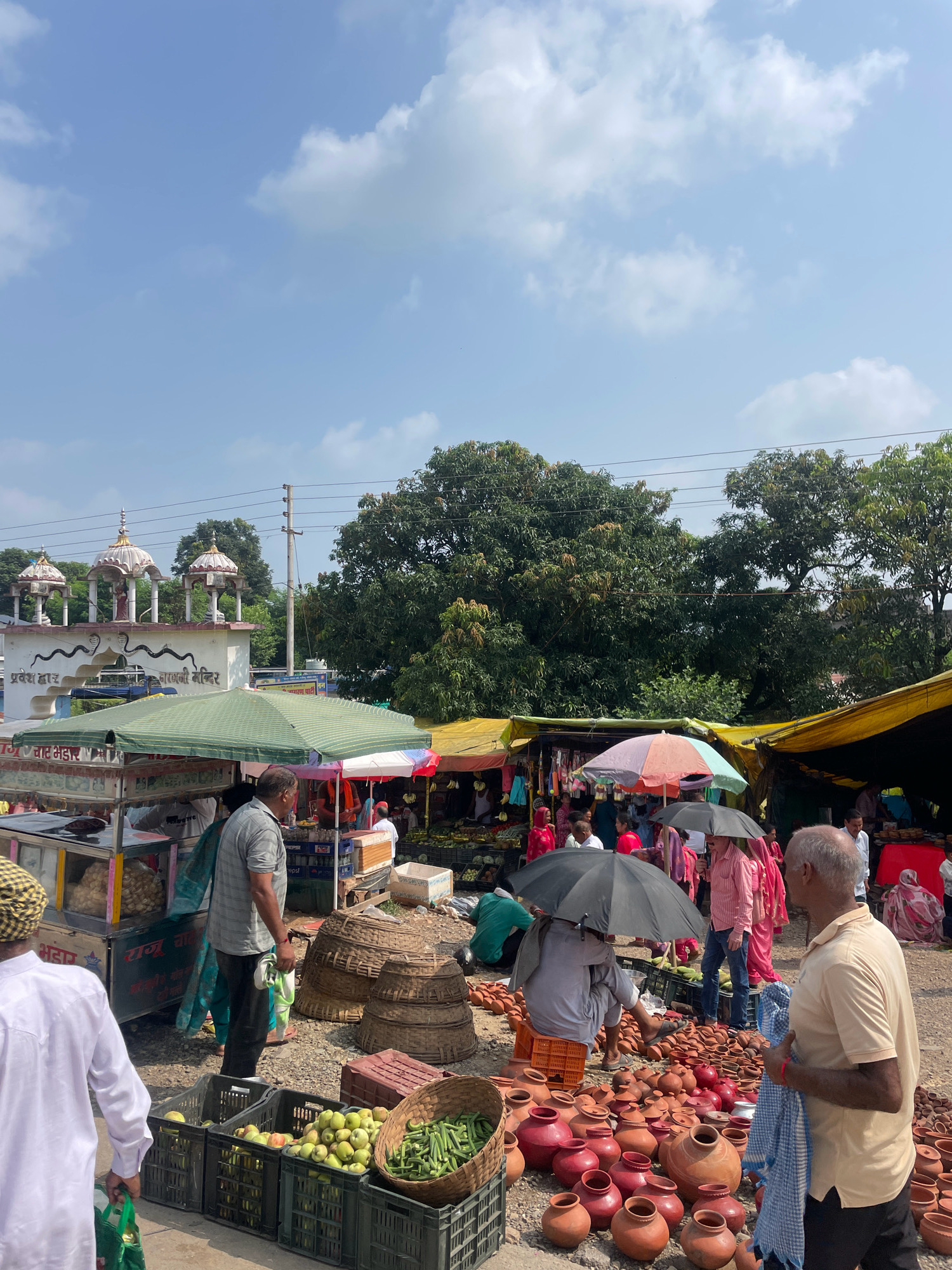This is a bustling outdoor market scene in Baud, India, likely situated near a temple, as indicated by the ornate white structure with domes and signage in Hindi. The market features numerous stalls, some covered with green and yellow tarps, selling a variety of goods. In the foreground, several crates of fresh green produce, including apples and beans, are displayed. Adjacent to the produce, numerous terracotta pots and clay vessels are laid out on the ground.
Many people are present, engaged in shopping and selling. Some individuals are under umbrellas, suggesting a sunny or potentially drizzly day. Several vendors appear to be tending to their stalls, while shoppers browse the offerings. Clothing colors vary, with some individuals wearing brightly colored traditional Indian attire. The ground of the market is dusty dirt. Power lines are visible strung between poles in the background. The overall atmosphere is lively and typical of a local Indian market.