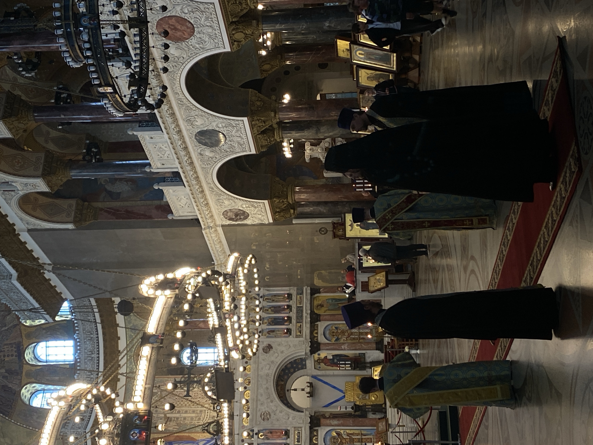 The image captures the interior of the St. Nicholas Naval Cathedral in Kronstadt, Russia, during what appears to be a religious service or ceremony. The grand hall features ornate architecture with high ceilings, arched colonnades, and intricate decorations. Large, multi-tiered chandeliers with numerous lights illuminate the space, casting a warm glow. Sunlight streams through high arched windows, creating atmospheric shafts of light, particularly noticeable in the central area of the image.
Several individuals in religious vestments are present, signifying the sacred nature of the event. On the left, a person in a blue and gold ornate robe stands facing away from the camera. In the foreground, two figures in dark robes, likely clergy, are seen. One is wearing a tall black headdress. In the mid-ground, more individuals in religious attire are positioned near an altar-like structure. Further in the background, other people, possibly worshippers or visitors, can be seen, some standing and some moving. Religious icons and paintings are displayed on stands and walls throughout the cathedral. The floor is polished, reflecting the light and the figures. A red carpet runner is laid out on the floor, leading towards the altar area. The overall impression is one of solemnity and grandeur within a historic and architecturally significant religious site.