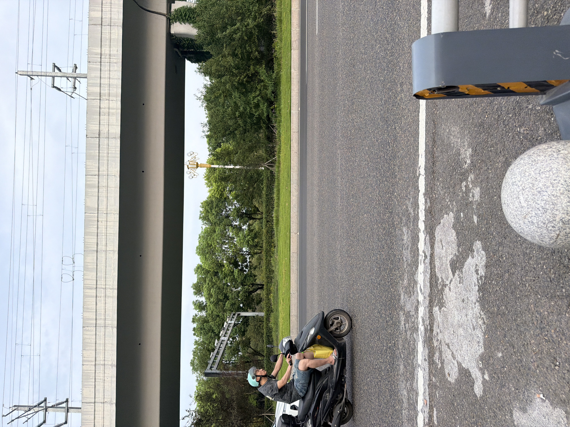 The image shows a person riding a black scooter on a road under an elevated concrete structure in Jiangjin, China. The rider is wearing a light blue helmet, a dark t-shirt, and denim shorts, with a yellow bag hanging from the scooter. The scooter is positioned in the right lane of a multi-lane asphalt road. In the background, there are green trees, a white pole with multiple lights, and overhead power lines. The elevated structure has a plain concrete surface with visible lines and some staining. The sky is overcast. The foreground shows the asphalt road with a white line, some wet patches, a granite sphere, and a grey barrier with yellow and black stripes.