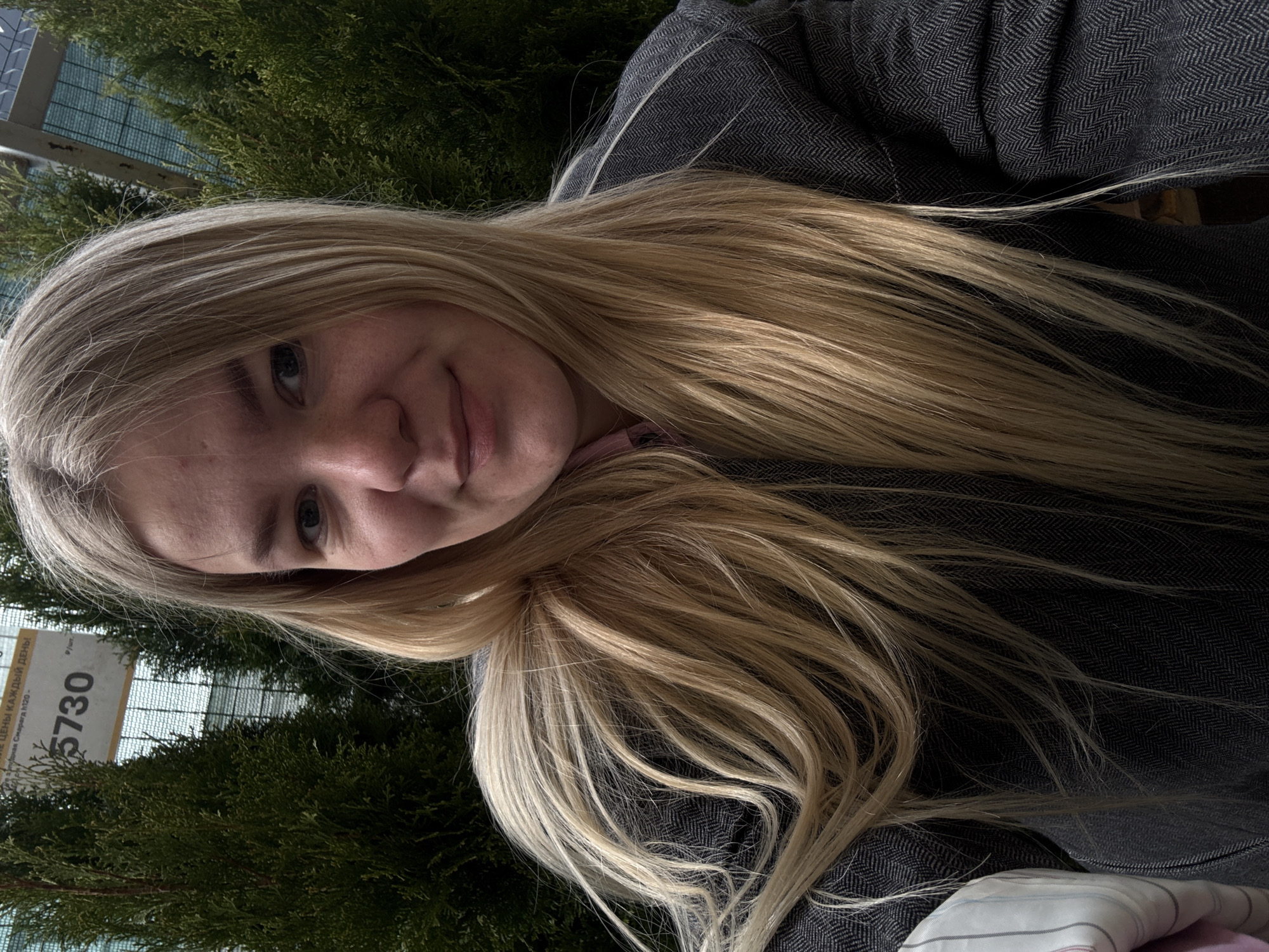A blonde-haired woman is photographed at an outdoor location in Izhevsk, Russia. The background includes coniferous trees, implying a park or garden setting. There's a sign with the number "5730" in Cyrillic lettering, partially visible. The woman is wearing a gray herringbone-patterned jacket. She is looking directly at the camera with a slight smile.