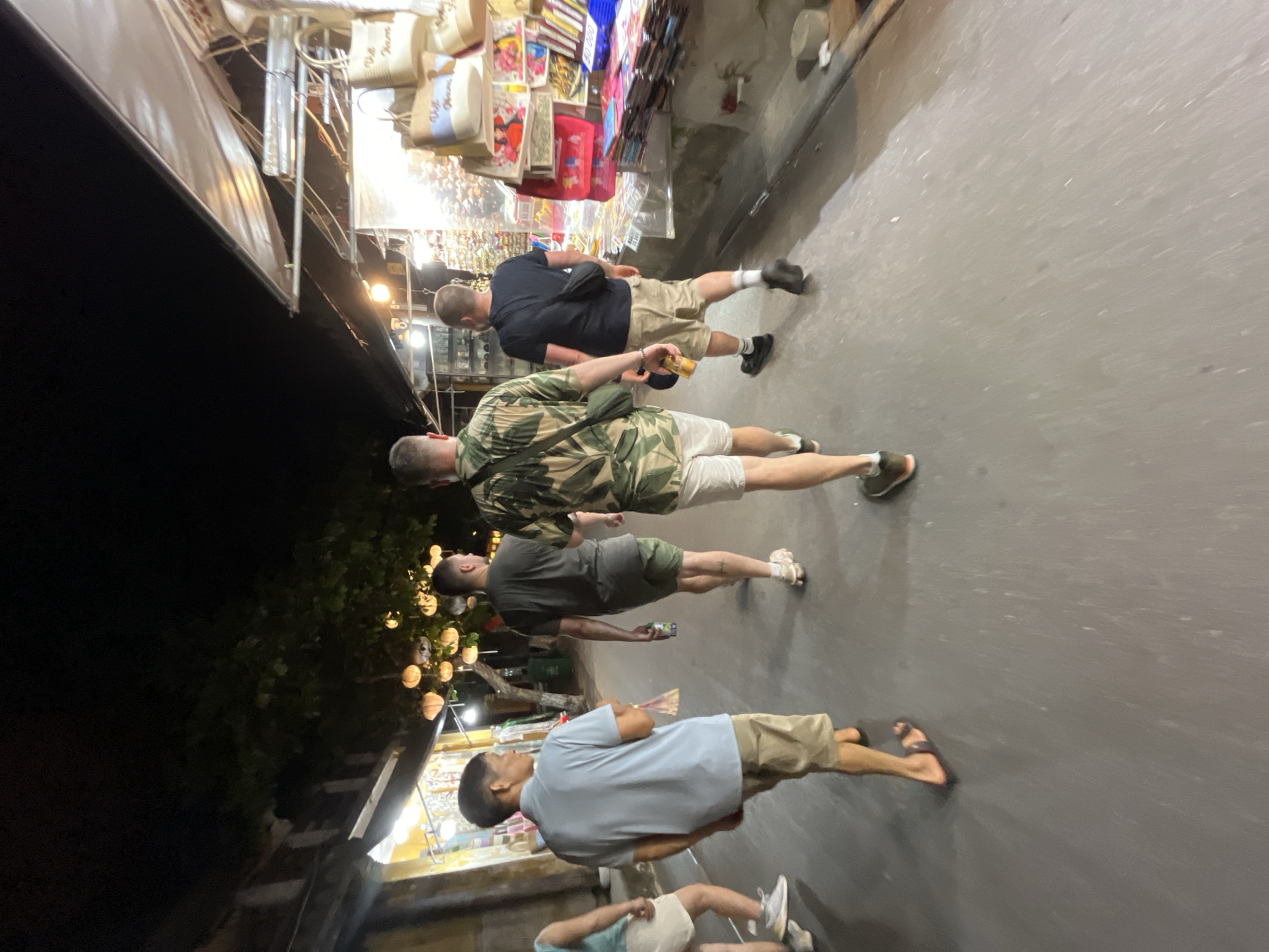 In Hội An Ward, Vietnam, four individuals walk down a street at night. The street is lined with shops displaying goods, and overhead, lanterns illuminate the scene. The pavement is dark and slightly wet, reflecting the ambient light. The individuals are walking away from the camera, their backs to us.
The person in the foreground to the left is wearing a light blue polo shirt and khaki shorts. Next to him, a man in a dark green t-shirt and olive green shorts walks with a bottle in his hand. In the center, a man in a green tropical-print shirt and white shorts leads the group, carrying a dark bag slung across his body. To the far right, a man in a black t-shirt and khaki shorts, also with a black bag, follows closely behind. The shop on the right displays an assortment of bags and colorful items, illuminated by bright lights. The shop on the left appears to be selling apparel and other merchandise. The overall atmosphere is that of a bustling night market.