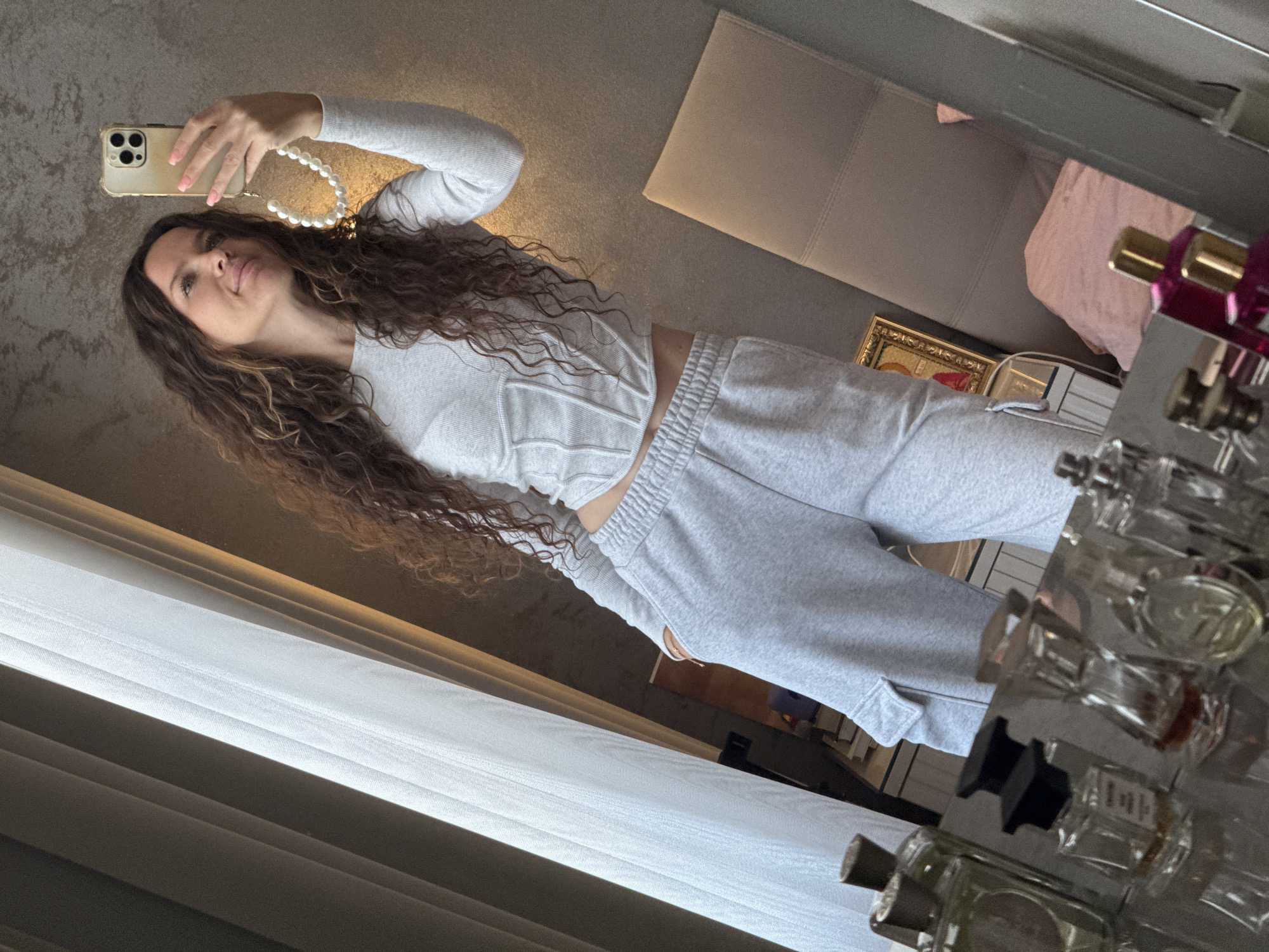 A woman with long, curly brown hair stands in front of a mirror, taking a selfie with her iPhone. She is wearing a light gray, long-sleeved cropped top with a corset-like detail and matching gray sweatpants with cargo pockets. Her left hand is in her pocket, and her right hand holds the phone, which has a pearl strap attached. The background shows a window with white sheer curtains on the left, and a bed with a pink pillow on the right. On a reflective surface in the foreground, several perfume bottles are visible. The scene appears to be indoors, possibly a bedroom, in an apartment in Khimki, Russia.