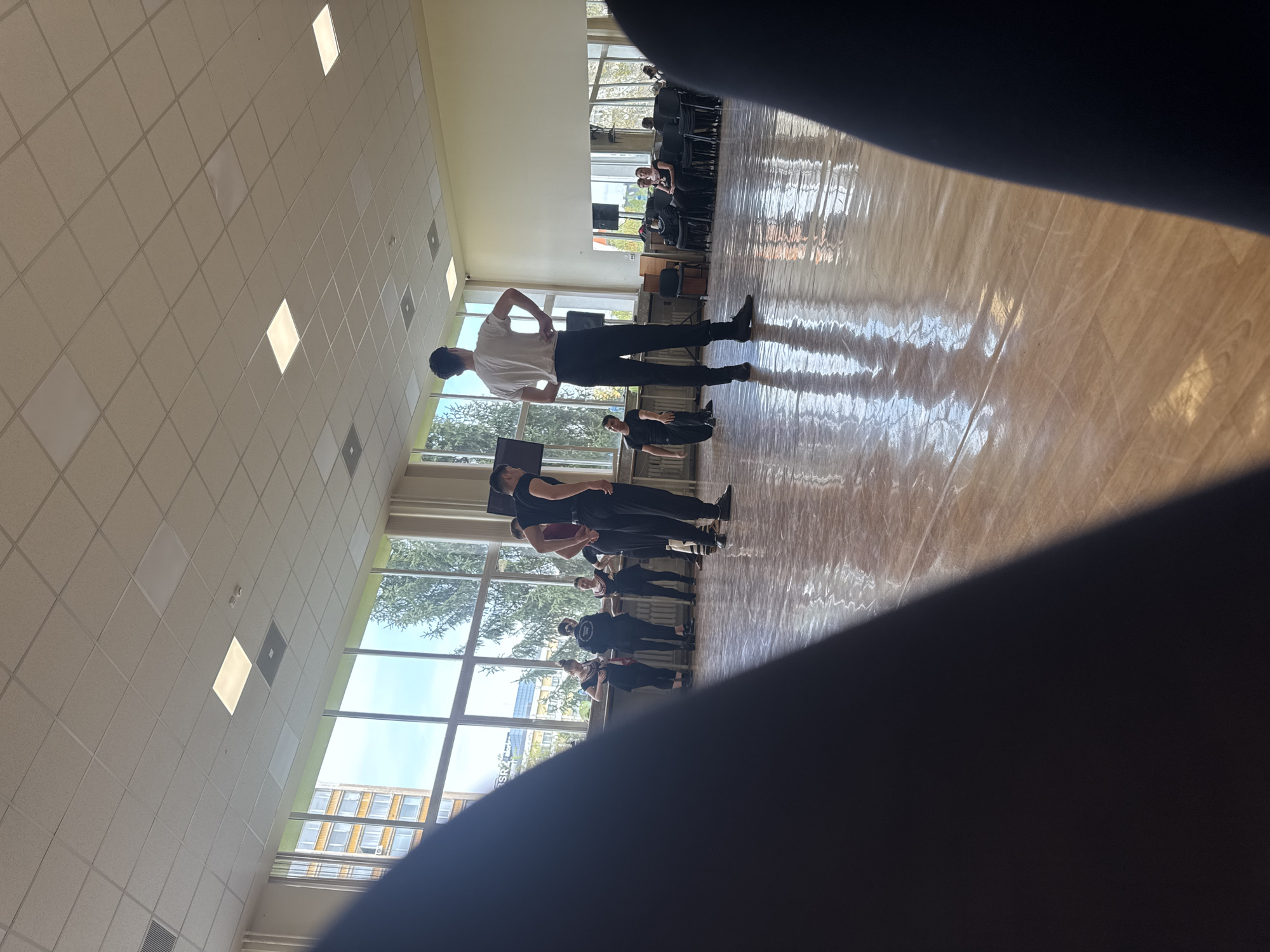 The image captures an indoor scene, likely a dance studio or a large multipurpose hall, featuring several individuals engaged in what appears to be a rehearsal or practice session. The room boasts a bright, reflective wooden floor and a high ceiling fitted with square acoustical tiles and evenly spaced rectangular light panels, which cast distinct reflections on the polished floor.
Numerous young adults are visible, mostly dressed in casual active wear like t-shirts and dark pants. In the foreground, facing away from the camera, a person in a white t-shirt and black trousers stands with hands on hips, striking a pose. Further back, other individuals are standing in various stances or formations, some seemingly observing or resting. In the far background, rows of stacked chairs suggest an area for spectators or waiting.
Large windows dominate the left side of the room, providing ample natural light and offering a view of the outside. Through these windows, green trees and an urban building with multiple windows are visible. The bright daylight streaming in, combined with the artificial ceiling lights, creates strong illumination, enhancing the reflections on the floor. A dark, out-of-focus area, possibly part of the camera operator's hand or clothing, obscures the lower right portion of the frame.
The visible text "SRZ" can be seen on a building or sign outside the window. Given the location context, this is a bustling indoor environment, potentially in Sofia, Bulgaria, with a focus on a group activity or performance. The time of day appears to be daytime due to the bright natural light.