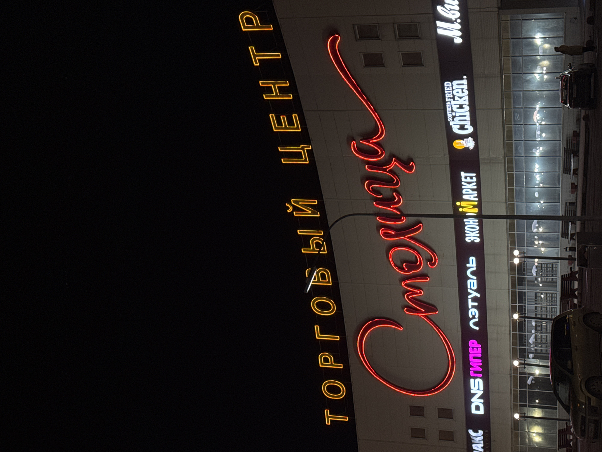 The image shows the facade of the Stolitsa shopping center in Perm, Russia, at night. Brightly lit vertical signs read "Торговый центр" (Shopping Center) in yellow and the name "Столица" (Stolitsa) in red neon. Other signs indicate stores and restaurants within the center, including DNS гипер (DNS Hyper), Л’Этуаль (L'Etoile), Экомаркет (Ekomarket), Southern Fried Chicken, and M.Видео (M.Video). A car is parked near the entrance. Several street lights line the sidewalk. The building's glass facade is visible in the background.