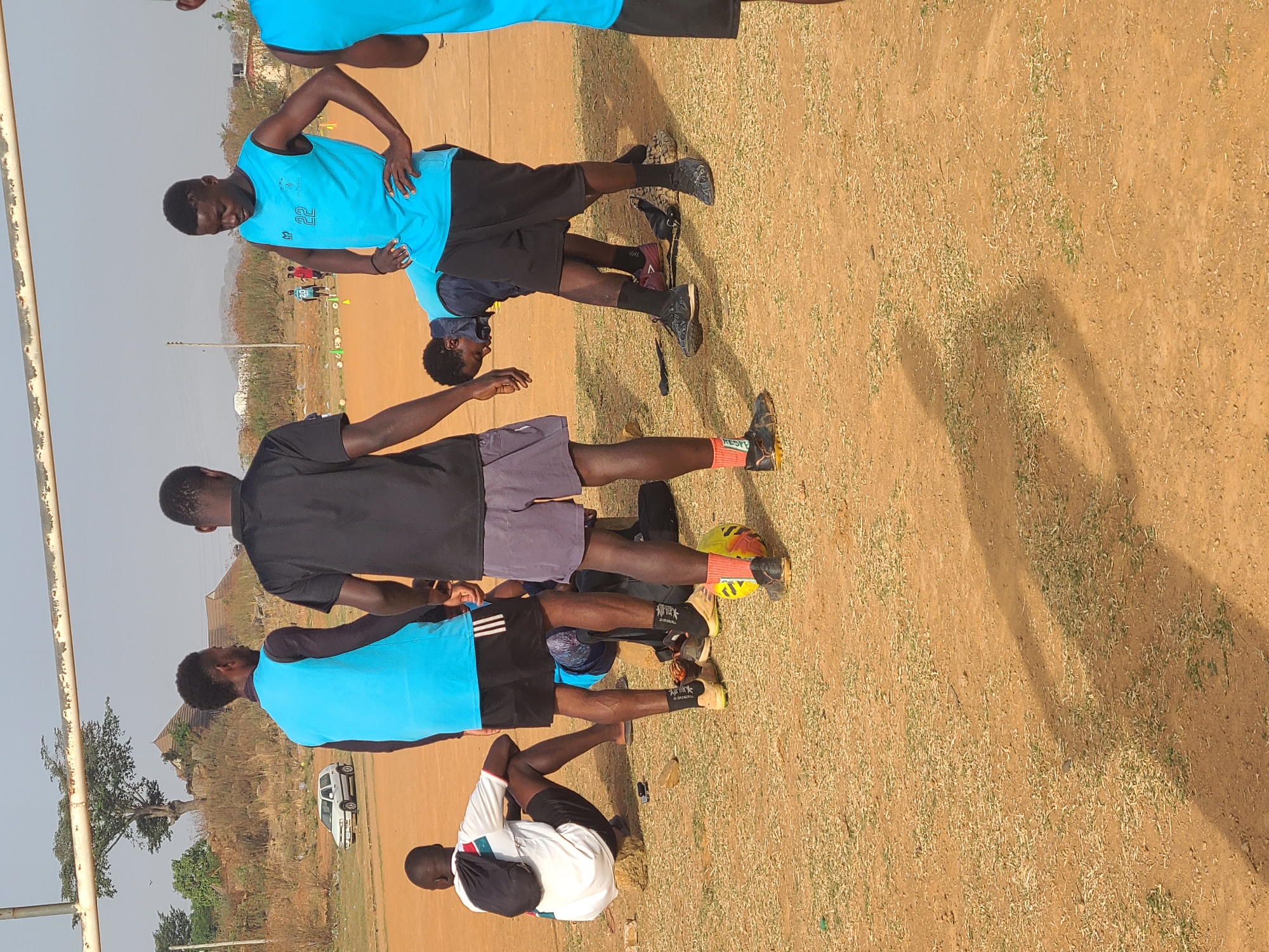 Several people stand on a dirt field in Barikin Jirga Summa, Nigeria. Some individuals wear blue athletic shirts and black shorts, while others wear black shirts and grey shorts. One person wears a white athletic shirt and black shorts with a backpack. They appear to be stretching or preparing for physical activity. The field is sparsely covered with vegetation. In the background, trees, a vehicle, and buildings are visible.