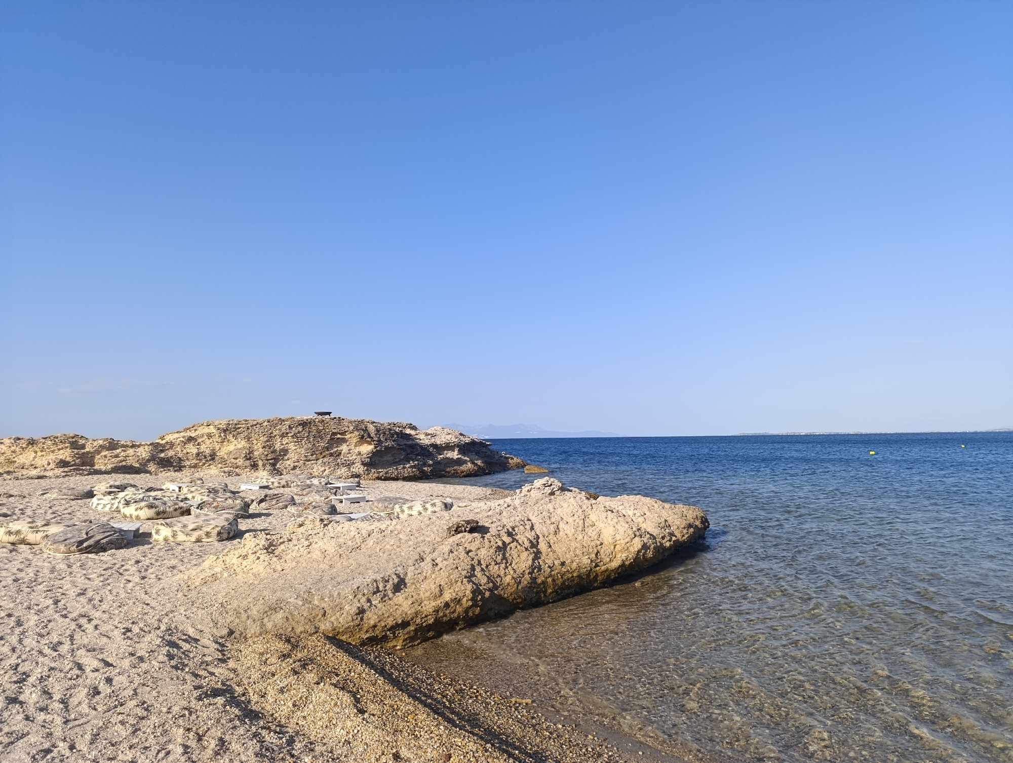The image depicts a serene beach scene in Fanari, Greece, under a clear blue sky. The foreground is dominated by a sandy beach with scattered rocks and pebbles. A large, tan-colored rock formation extends into the clear, calm sea on the right side of the image. The water is shallow near the shore, allowing a view of the rocky seabed.
In the mid-ground, more rocky outcrops rise from the sand, with several large, cushioned loungers and low white tables arranged on the beach, suggesting a seating area. These are set against a backdrop of the rocky terrain.
The background features the vast blue expanse of the Aegean Sea, meeting the horizon where a distant, faint landmass is visible. The water is deep blue, with a gentle ripple. Two yellow buoys are visible in the distance, marking the sea. The overall atmosphere is peaceful and tranquil, characteristic of a quiet coastal resort area.