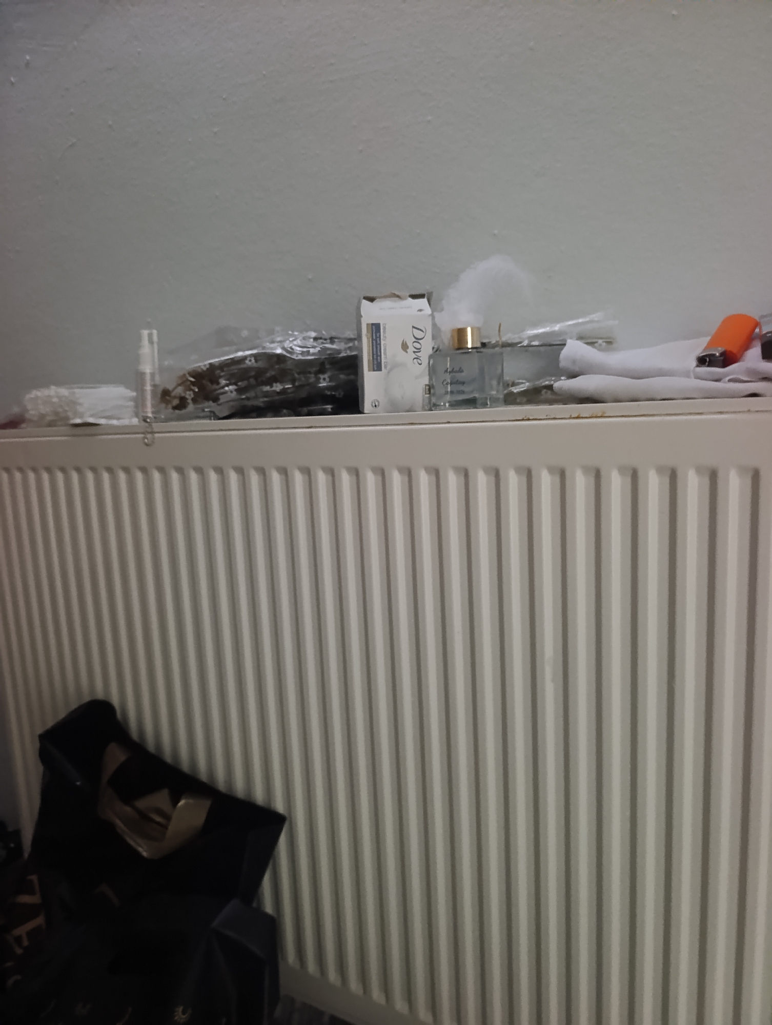 A shelf above a white radiator holds several items. On the left, a small clear bottle with a silver cap is visible, followed by a pile of what appears to be individually wrapped small items. Next to this is a box of Dove soap, a glass bottle with a gold cap containing a light blue liquid, and a folded white cloth. To the right of the cloth, an orange lighter rests on top of another folded white cloth. The radiator is a standard white model with vertical fins. In the lower left of the image, a black tote bag is partially visible. The background is a plain, light-colored wall. The scene is indoors, likely in a residential setting.