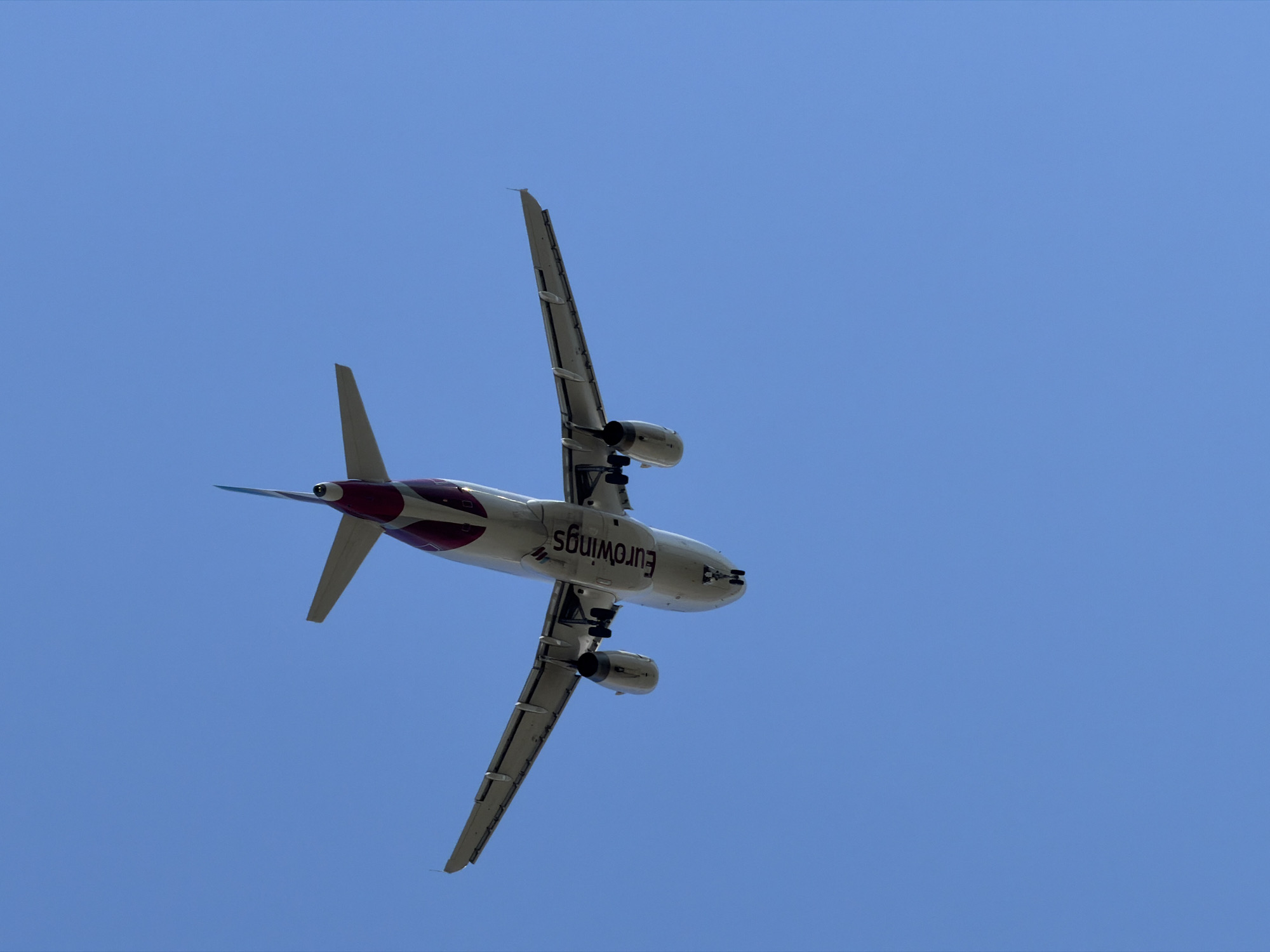 An airplane, identified as a Eurowings aircraft, is captured from a low-angle perspective, appearing to be in flight against a clear blue sky. The underside of the plane is visible, revealing its wings, engines, and tail. The "Eurowings" logo is clearly discernible on the fuselage. The landing gear is deployed, suggesting the aircraft may be preparing to land or has just taken off. The image does not depict any specific urban or natural landmarks that would pinpoint its location within the city of Burgas, Bulgaria.