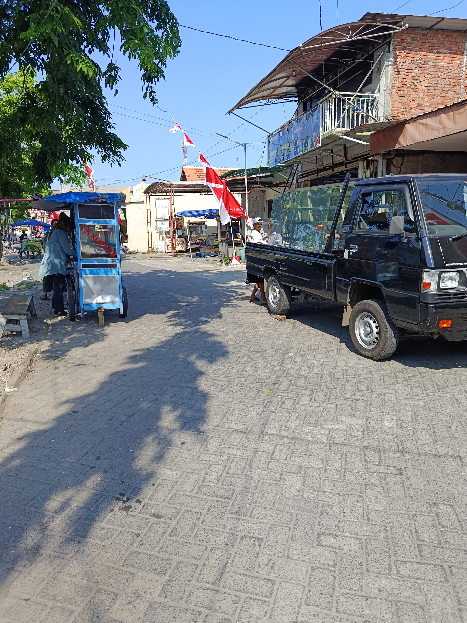 The image depicts an outdoor scene in Keboansikep, Indonesia, on a sunny day with a clear blue sky. A street paved with interlocking bricks is visible, with shadows of trees and people cast across it. On the left, a blue food cart with a glass display case and a blue tarp is parked. A person wearing a light blue shirt and dark pants stands behind the cart, facing away from the camera. Adjacent to the cart, a wooden bench is placed on the side of the road.
In the center and right of the image, a black pickup truck is parked. The truck has its doors open, and two individuals are visible inside the cab. One person, wearing a white cap, is looking out the window. Another person is also in the cab. The truck's rear bed is partially visible, with a glass enclosure. Red and white Indonesian flags are displayed on poles in the background, suggesting a national celebration or event. Behind the truck, a two-story building with a brick facade and a corrugated metal awning is situated. Other smaller food stalls with blue awnings are also visible in the background. The overall atmosphere suggests a bustling street market or roadside commercial area.