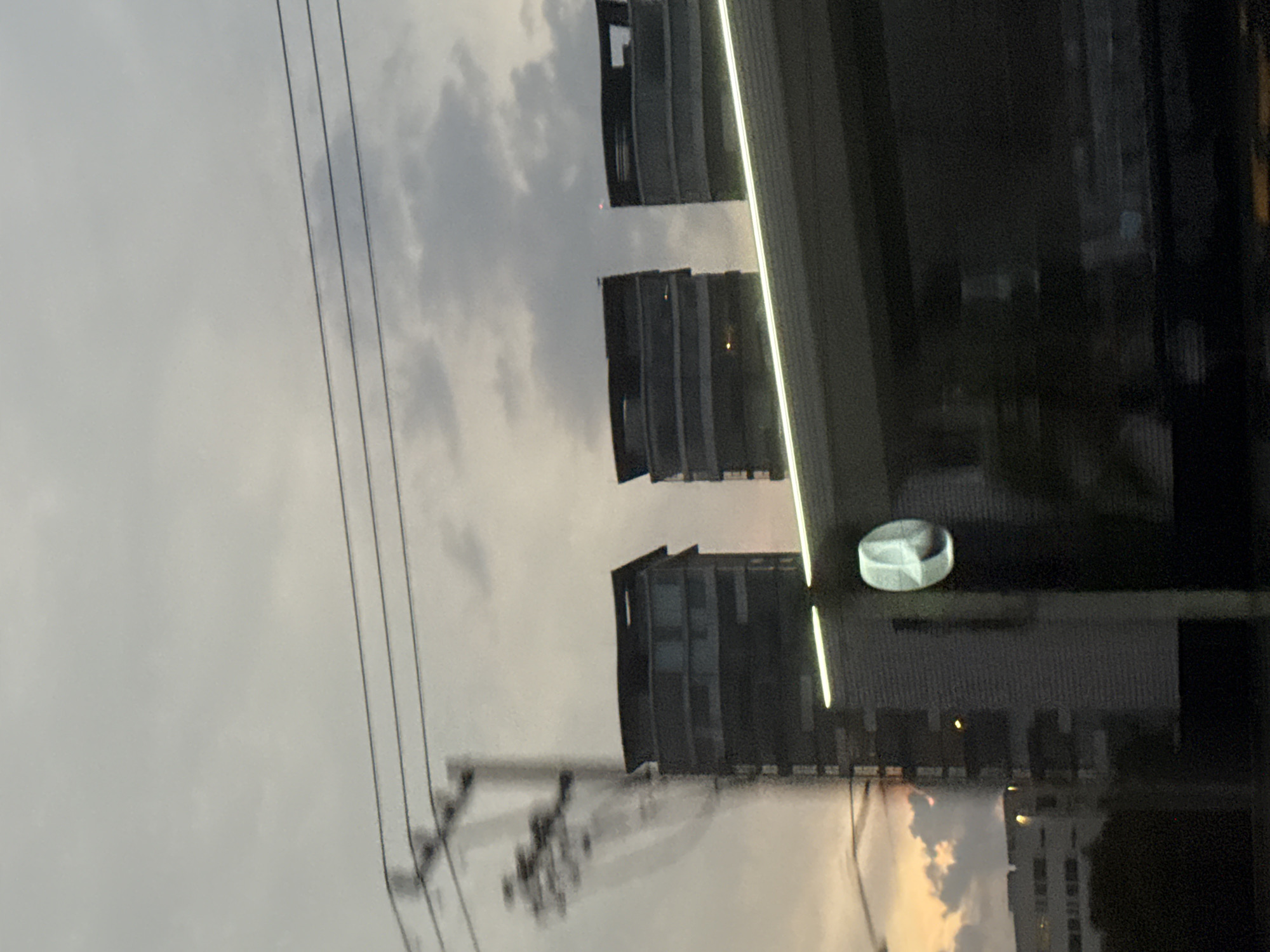 The image shows a view of buildings, power lines, and a cloudy sky. The buildings are multi-story structures, and the power lines are strung across the sky. A reflective surface (likely a window) creates a double exposure, showing both the exterior scene and a reflection of the interior. The scene appears to be taken in an urban or suburban area, potentially in San Bernardino Tlaxcalancingo, Mexico. A round object is visible near the reflective surface, its identity is unclear. No people are visible.