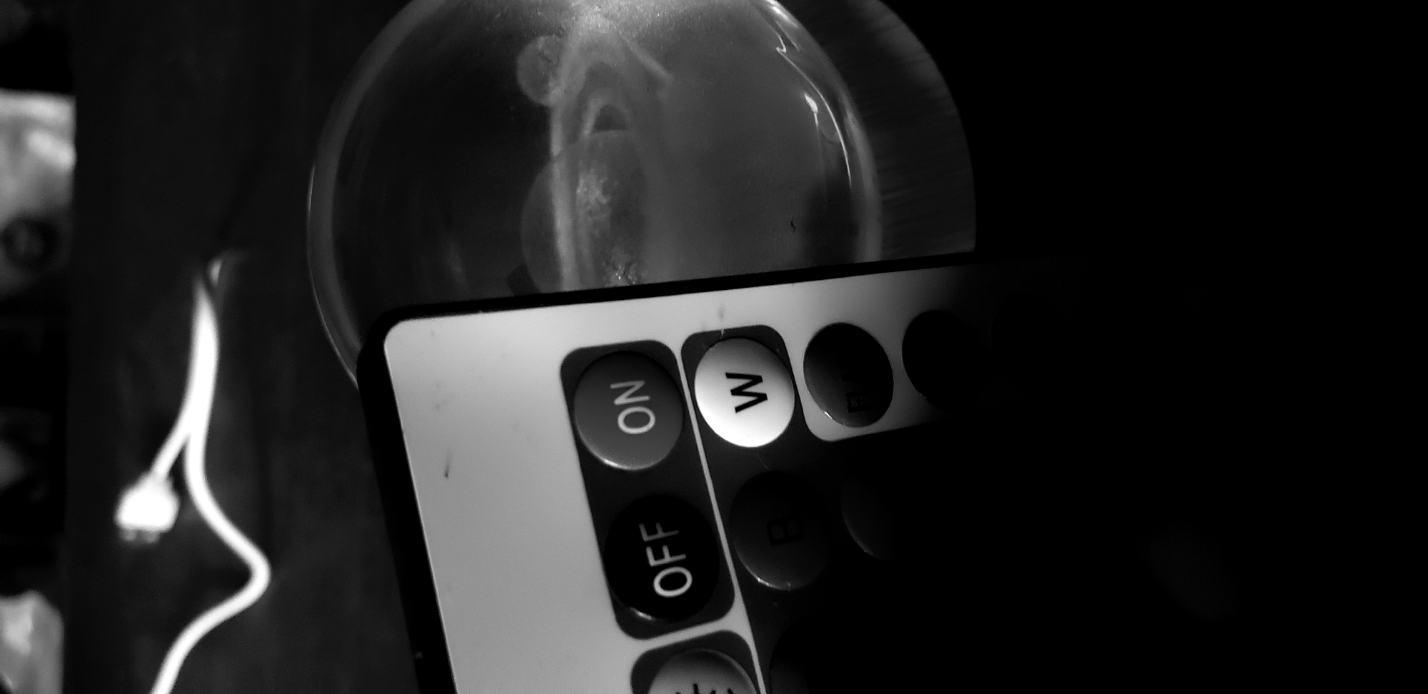 The image shows a close-up of a remote control positioned in front of a lightbulb. The remote features buttons labeled "ON", "OFF", and "W". A power cord is visible to the left of the lightbulb. The scene is in grayscale and has low lighting. The location in Narayi, Nigeria is unspecified.