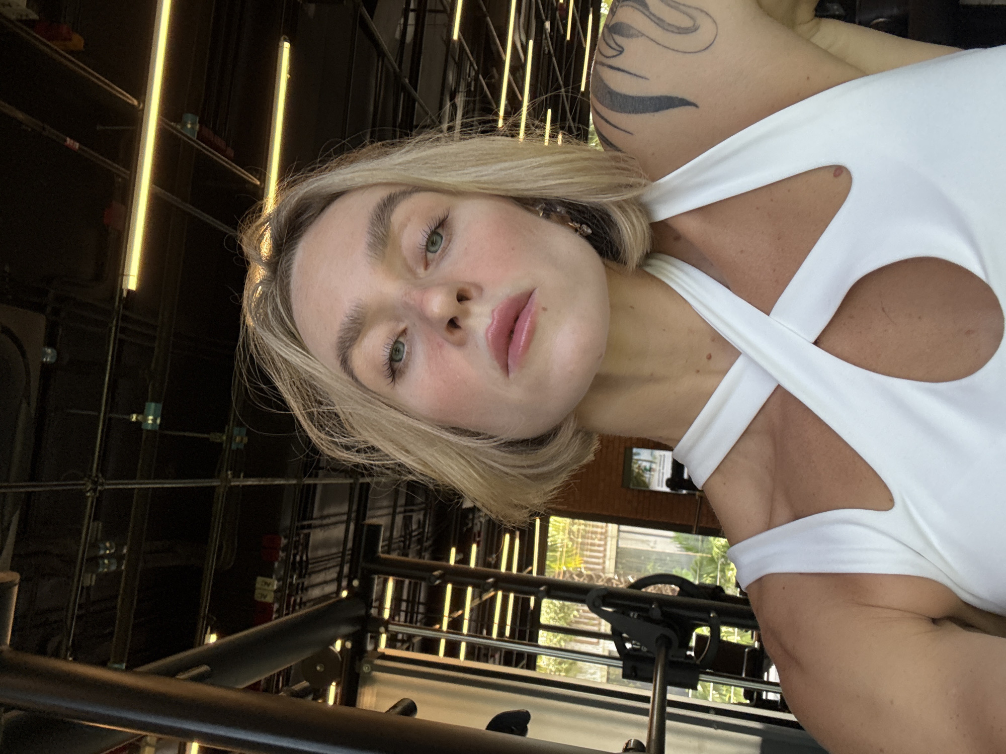 A medium shot captures a blonde woman with light makeup, wearing a white halter top. She is looking directly at the camera, with her head slightly tilted. Her short blonde hair frames her face, and her eyes are a striking blue-green. The background shows the interior of a gym, with metal equipment, exposed wiring, and fluorescent lights visible on the ceiling. Natural light streams in from windows in the background, revealing foliage and trees outside, suggesting a tropical location. The overall impression is a selfie taken at a fitness center in Rawai, Thailand.