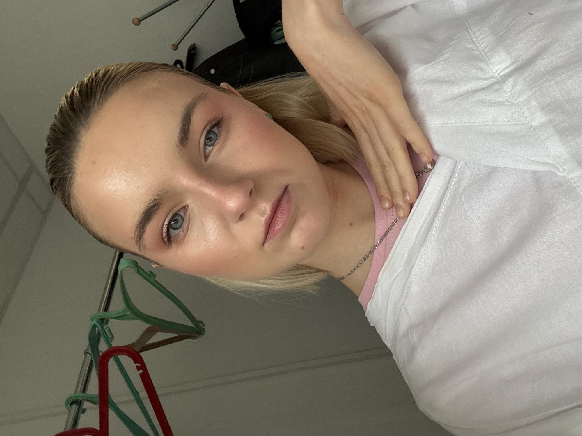 A close-up, angled portrait of a young woman with fair skin, blonde hair styled back, and light blue eyes. She is wearing a white button-down shirt over a pink top. A silver necklace is visible around her neck. Her right hand is resting near her collarbone. In the background, a clothing rack with colorful hangers is visible. The room appears to be a simple domestic interior. The photograph does not provide enough information to determine a specific location within Saint Petersburg, Russia.