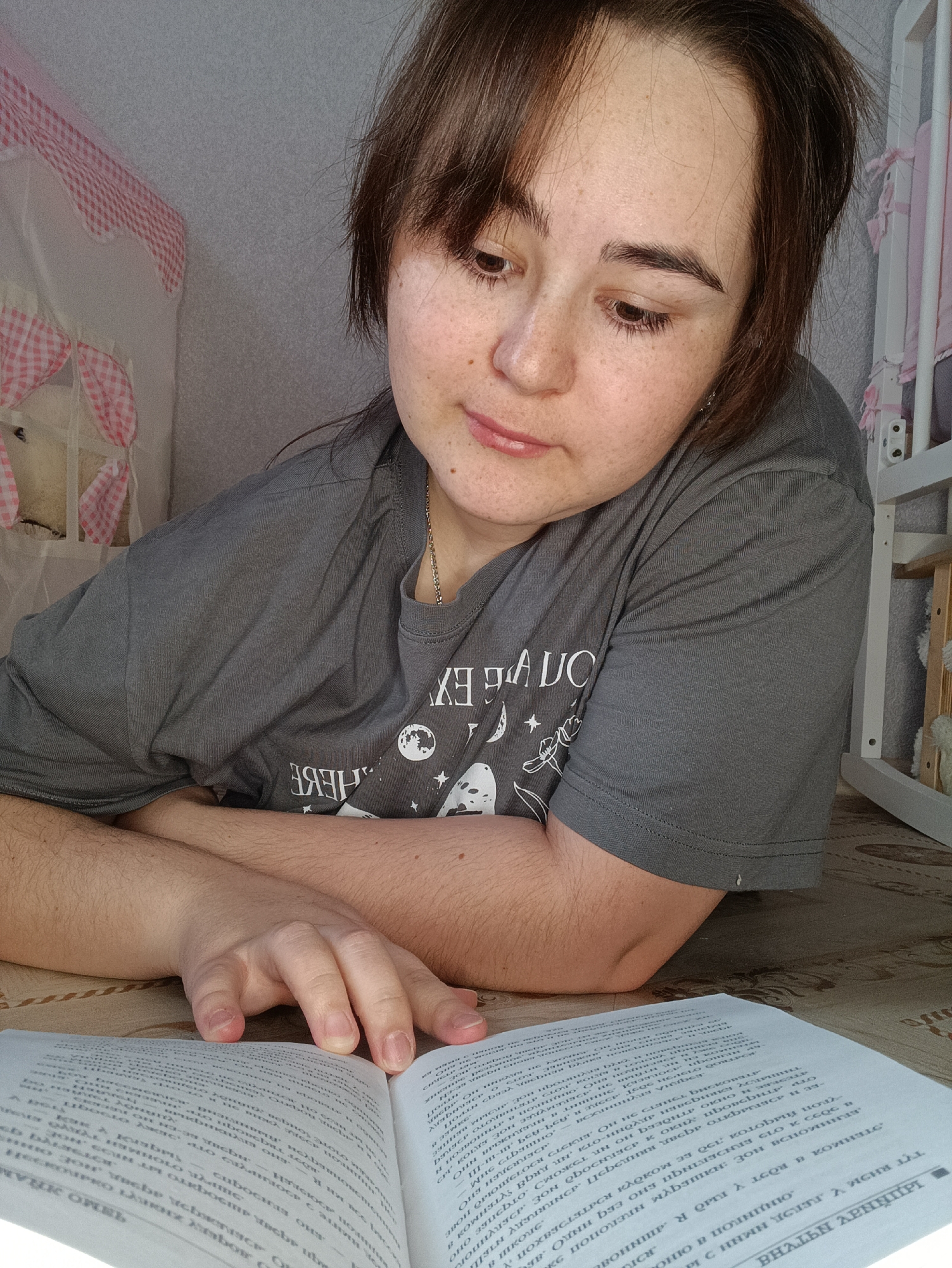 A woman with brown hair and freckles is lying on the floor, reading a book written in Russian Cyrillic. She is wearing a grey t-shirt with a moon and star design. A baby's crib with a pink and white gingham pattern is visible in the background. The location is unspecified.