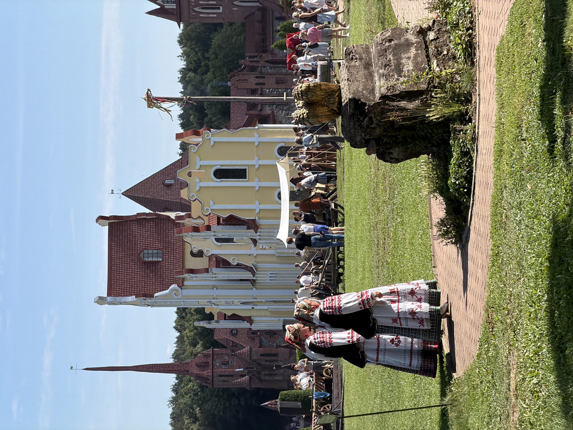 The image depicts a historical reenactment or festival taking place in a park-like setting with a distinct architectural style. In the foreground, two individuals dressed in traditional Belarusian folk costumes, featuring white embroidered dresses and headscarves, are walking on a brick pathway. Behind them, a crowd of people, some in period clothing and others in modern attire, are milling around in a grassy area. Several tents and market stalls are visible, suggesting a fair or event. In the background, elaborate buildings with red tiled roofs and white decorative elements stand out. One building has a tall, slender spire, reminiscent of a church or castle tower. The overall environment is bright and sunny, with lush green grass and trees in the distance. The presence of traditional costumes and historical-looking architecture strongly suggests a cultural event in a place that values its heritage, such as the Літвенскі сельскі Савет in Belarus.