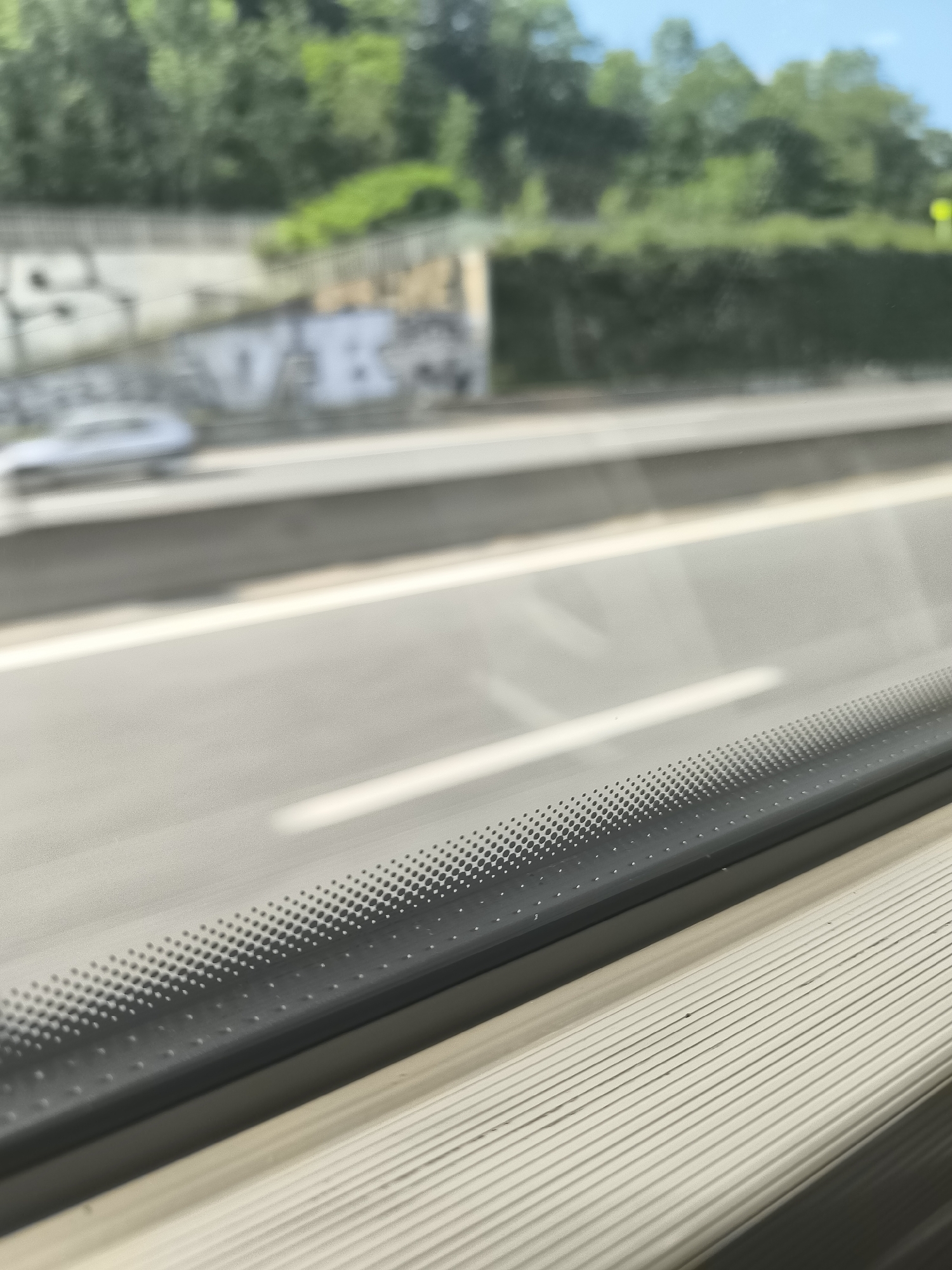 The image shows a view from inside a moving vehicle, likely a train or bus, looking out onto a highway. The foreground contains the interior window ledge of the vehicle, with a ridged, off-white surface and a black, perforated window frame. The background, seen through the window, is a highway with a white dividing line. Beyond the highway is a concrete wall with graffiti, followed by a slope covered with green trees and bushes. Based on prior context clues, the location is presumed to be Saint-Chamond, France.