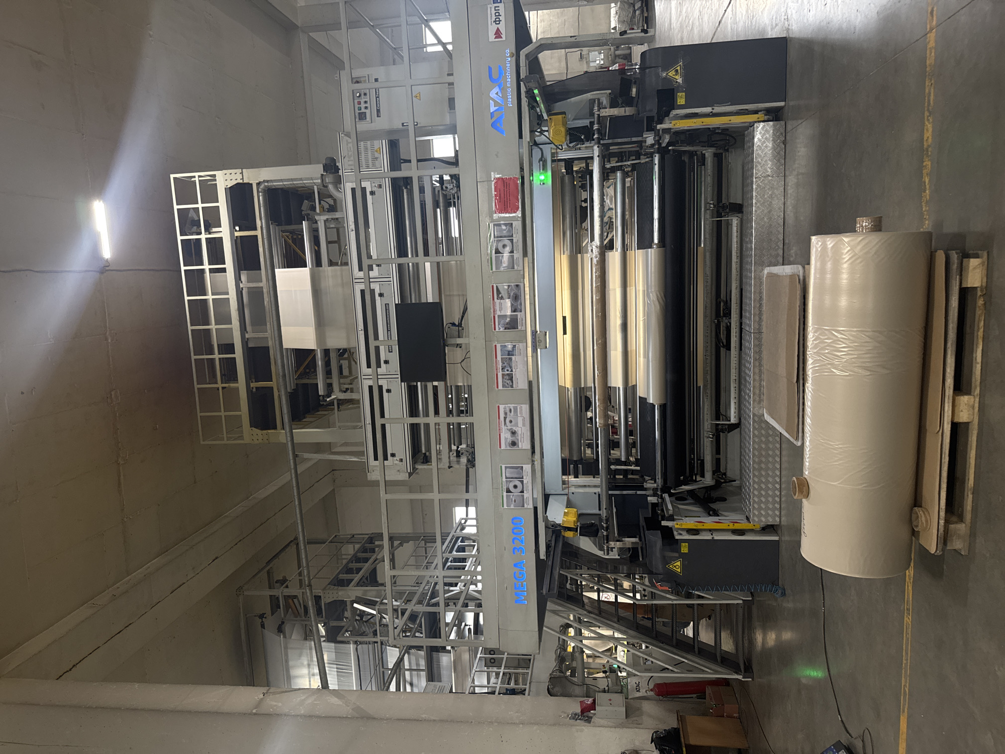 The image captures the interior of an industrial facility in Dikaya Gar', Russia, housing a large, multi-story plastic film extrusion or lamination machine. The machine, branded "MEGA 3200" and "ATAC plastic machinery co.," is actively processing translucent, light brown plastic film, which is threaded through numerous rollers and winding mechanisms across multiple levels of the apparatus. A monitor is mounted on the front-facing section of the machine, below signs indicating "Corona Treat" and "LÜCET."

Large rolls of the finished plastic film, wrapped around cardboard cores, are visible within the machine's winding section and also stacked on a wooden pallet on the concrete floor in the foreground. The facility features a high, unpainted concrete ceiling exhibiting cracks, supported by large concrete columns and beams. Industrial fluorescent tube lighting illuminates the space, with one fixture emitting a bright, focused beam. The concrete floor has visible yellow demarcation lines. A red fire extinguisher is positioned on the left side near the base of the machine. Metal stairs and platforms are integrated into the machine's structure, allowing access to its upper sections. No individuals are present in the scene.