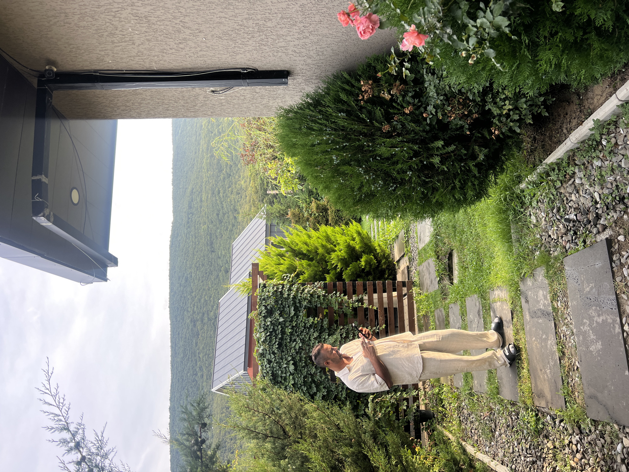 A man is standing on a stone pathway in a garden. He is wearing a beige linen outfit and black sandals. The man is looking at his phone. The garden has several trees and bushes, as well as a wooden fence covered in ivy. In the background, there are two houses with dark gray roofs and a hill covered in trees. The sky is overcast. The location is in Tsodoreti, Georgia.