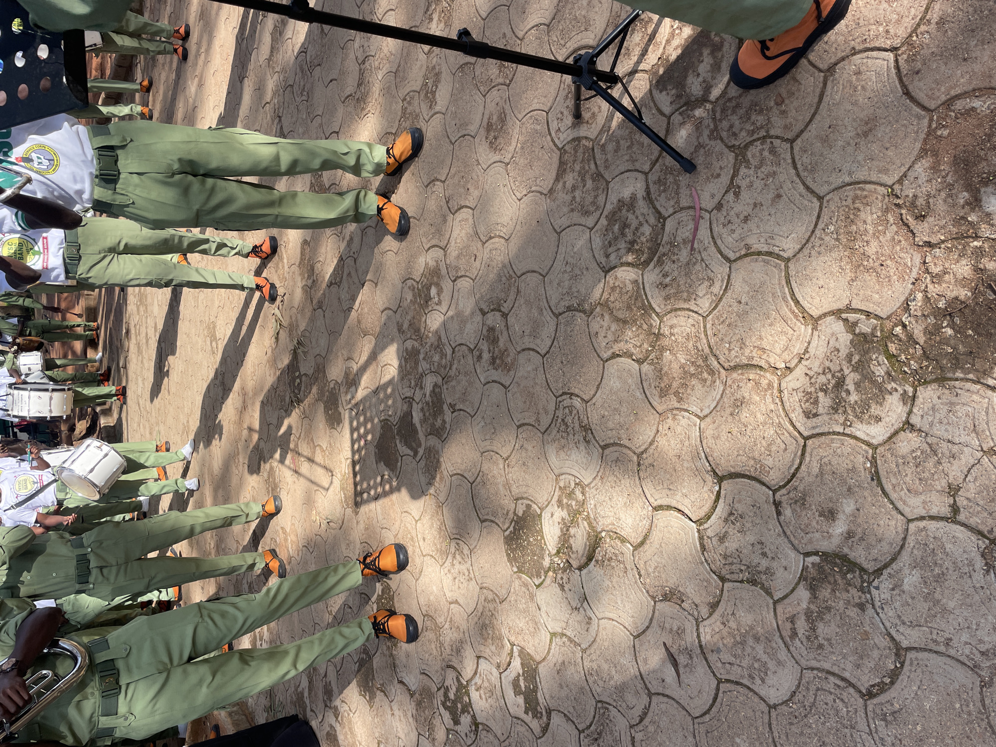 The image depicts a group of individuals in green uniforms, some holding musical instruments, standing on a paved outdoor surface. The pavement is composed of interlocking grey concrete tiles with a textured, possibly geometric, pattern. The scene appears to be outdoors, with sunlight casting shadows of the people and a tripod onto the ground.
Specifically, the individuals are wearing olive green trousers and matching shirts with dark green belts. Some are also wearing white t-shirts underneath their uniforms. Several people are carrying brass instruments, and at least one person is holding a white drum with a strap. The footwear visible consists of bright orange shoes with black soles and laces.
In the background, other individuals in similar green uniforms are visible, some holding drums. The overall impression is that of a band or marching group. The environment suggests a public space, possibly a parade ground or a school compound in Jos, Nigeria, given the context provided. The ground is somewhat worn and shows signs of dirt and debris in the crevices between the tiles. A black tripod, likely for a camera or lighting, is positioned to the right of the frame.