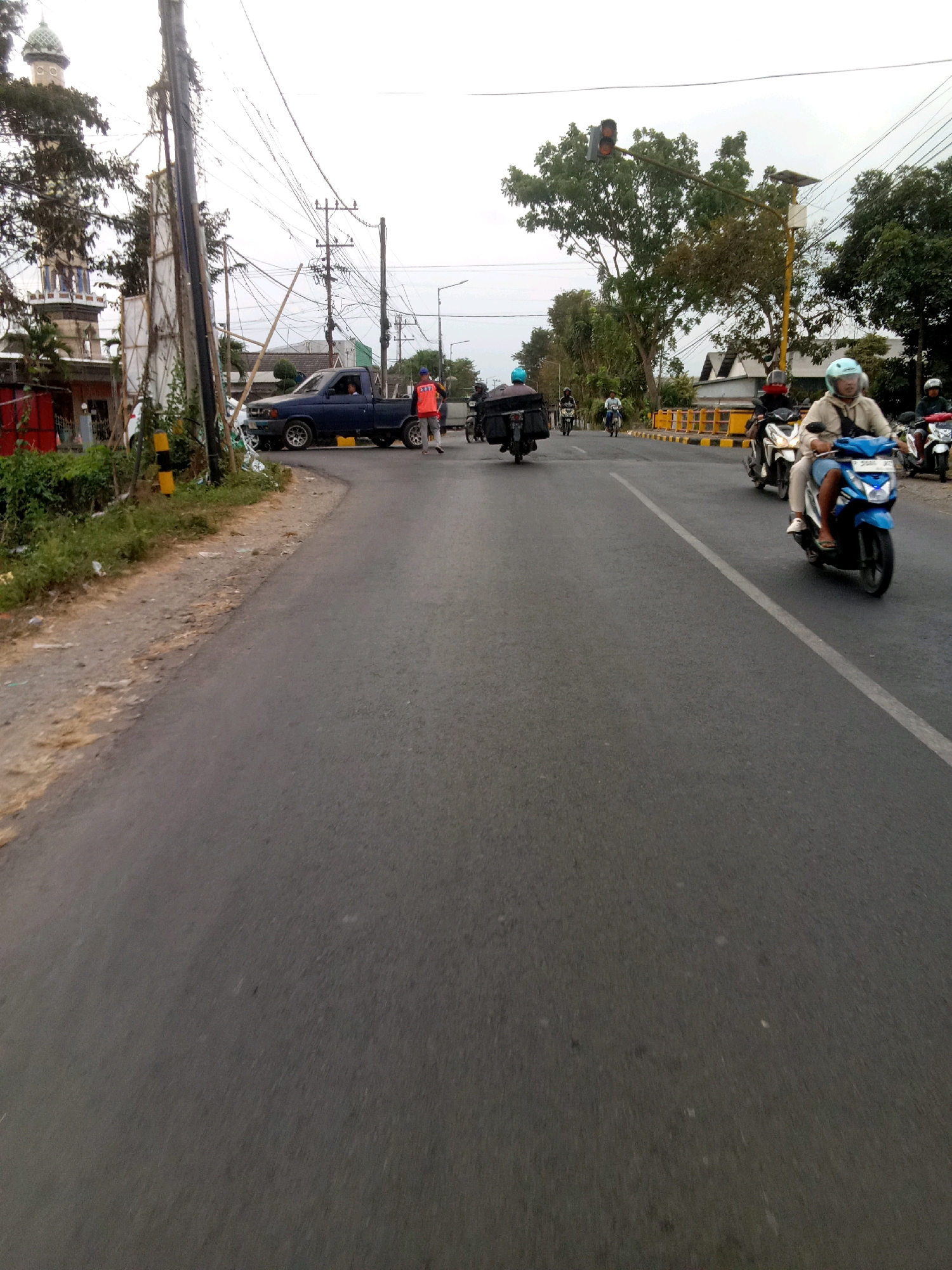 The image shows a street scene in Pancakarya, Indonesia. In the foreground, the asphalt road is visible with a white lane marking. Several motorcycles are on the road, with riders wearing helmets. One motorcycle in the right middle ground is blue, and the rider is wearing a light-colored jacket and blue shorts. Further down the road, other motorcycles are also visible. On the left side of the road, there is a blue pickup truck with a person in the driver's seat. A man in an orange vest is standing near the truck. To the left of the road, there are trees, a mosque with a dome, and some buildings. Utility poles with numerous wires are present along the side of the road. A traffic light is visible in the distance, with the red light illuminated. The sky is overcast.