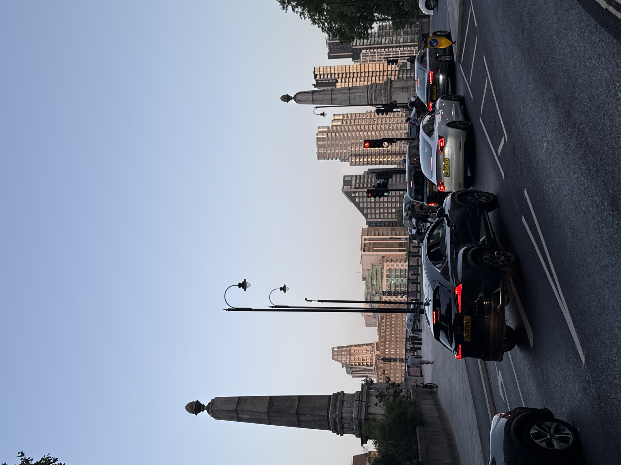 The image shows a street scene in London, United Kingdom, likely on a bridge, with a clear sky. In the foreground, there are several cars on a multi-lane road. A black Lexus SUV is in the left lane, followed by a white Tesla Model 3 in the center lane. To the right of the Tesla, a dark grey Audi sedan is also in the center lane. Further down the road, a black taxi is visible.
Along the side of the road, a paved walkway is present with a few pedestrians. On the left side of the walkway, a large, stone obelisk-like monument with an acorn-shaped finial stands prominently. Further down, there are two ornate lampposts with multiple lights.
In the background, the cityscape of London is visible, characterized by a cluster of modern high-rise buildings. The architecture suggests a contemporary urban environment. The lighting indicates it is either early morning or late afternoon, with the setting or rising sun casting a warm glow on some of the buildings. The overall atmosphere is that of a busy urban thoroughfare.