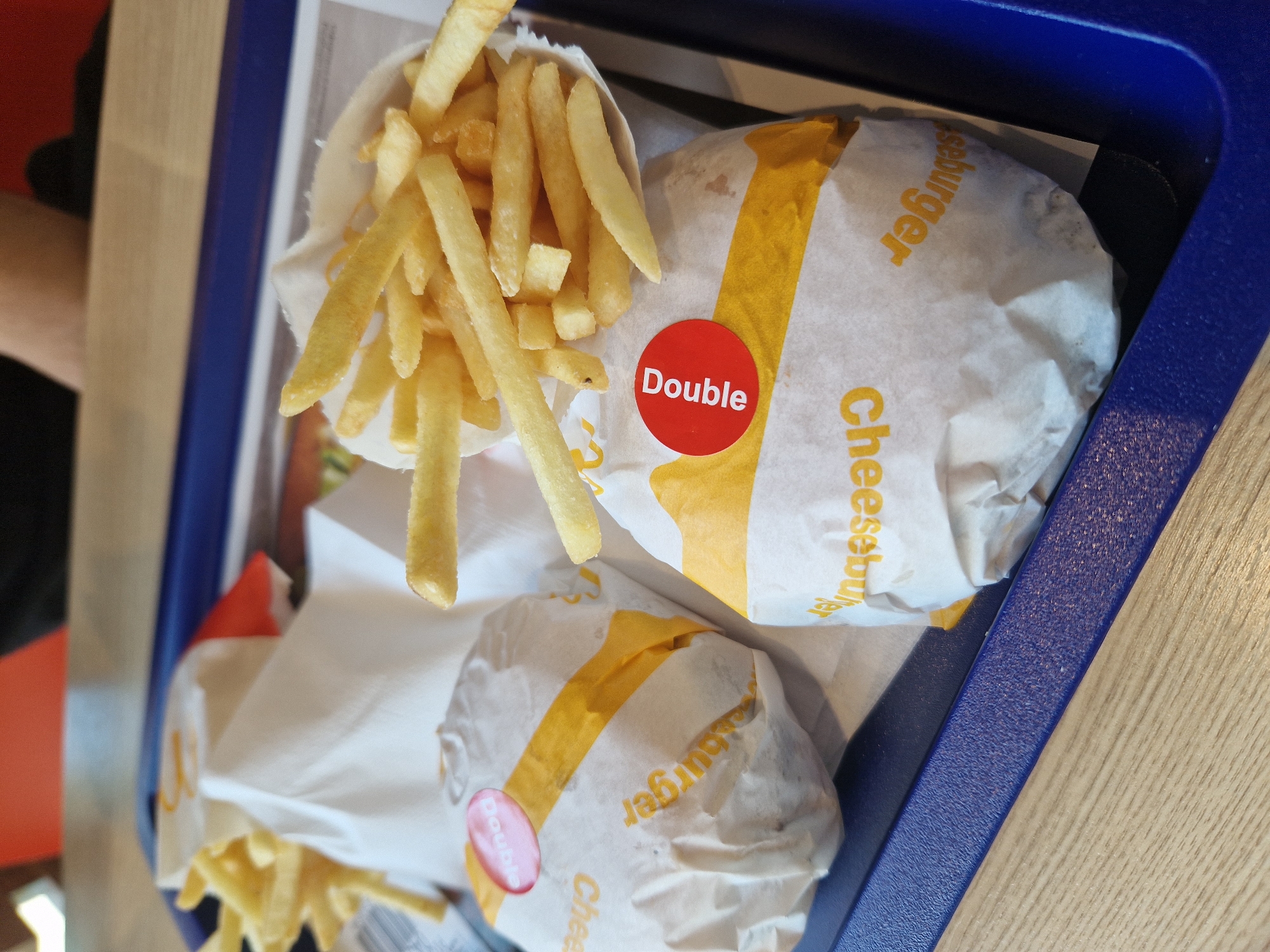 Two cheeseburgers, wrapped in white paper with yellow and orange branding, and a portion of french fries are arranged on a blue plastic tray. One burger is on the left and slightly further back, while the other is on the right and closer to the viewer. The burger on the right has a red circular sticker with the word "Double" in white lettering. The french fries are in a white paper container, and some are spilling out onto the tray. The tray is placed on a wooden surface.