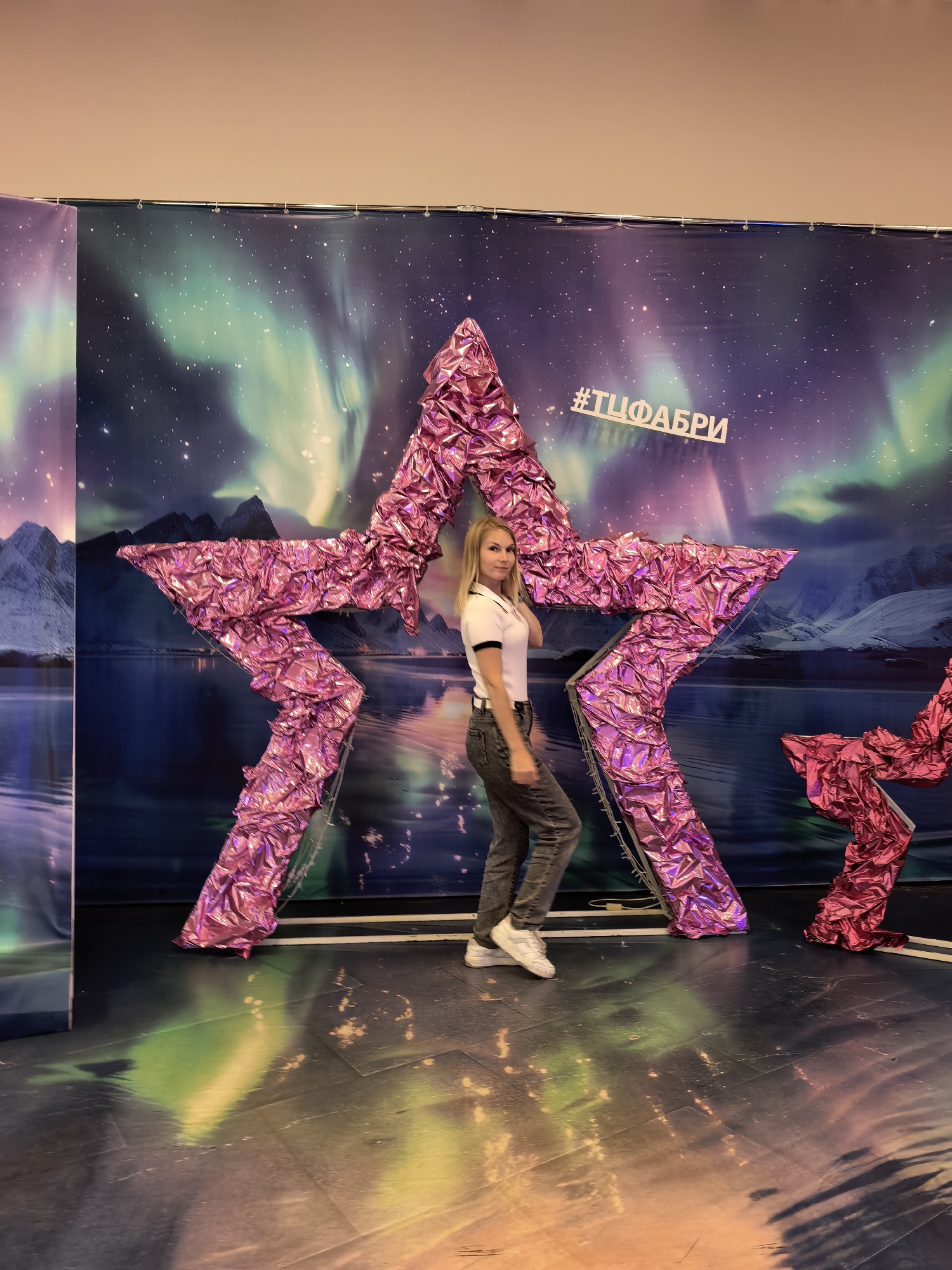 A full-body, slightly low-angle shot shows a young woman posing in front of a large, pink, foil-covered star decoration. She is standing to the left of the star, facing the camera with her right leg forward and her left arm resting on her hip. The woman has blonde hair and is wearing a white polo shirt with a black collar and trim, dark gray jeans, and white sneakers. The star is a prominent, three-dimensional object made of crumpled pink foil, with a metal frame visible underneath. Behind the star is a large backdrop depicting a night sky with the aurora borealis, mountains, and a body of water. The hashtag "#ТЦФАБРИ" is visible in white text on the backdrop. The floor in front of the decoration is dark and reflective, showing colorful light patterns from the backdrop and the star. The scene appears to be an indoor event or photo opportunity, possibly at a shopping center or exhibition in Sterlitamak, Russia, given the hashtag.