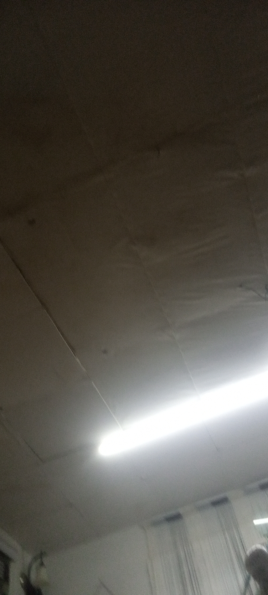 The image shows the interior of a room, seemingly lit by a fluorescent tube light. The ceiling is composed of rectangular panels. Below the light, there are translucent, hanging curtains or drapes and blurred figures. The location is Unknown.