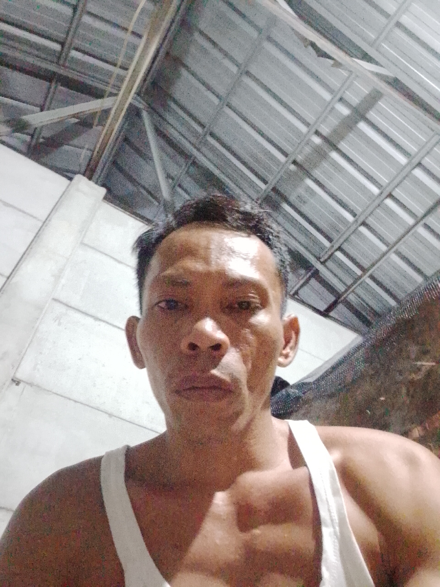 A man in a white tank top is facing the camera. The background consists of a white concrete wall on the left and a corrugated metal roof structure overhead. Based on the prompt, the location is Bukit Ketok, Indonesia.