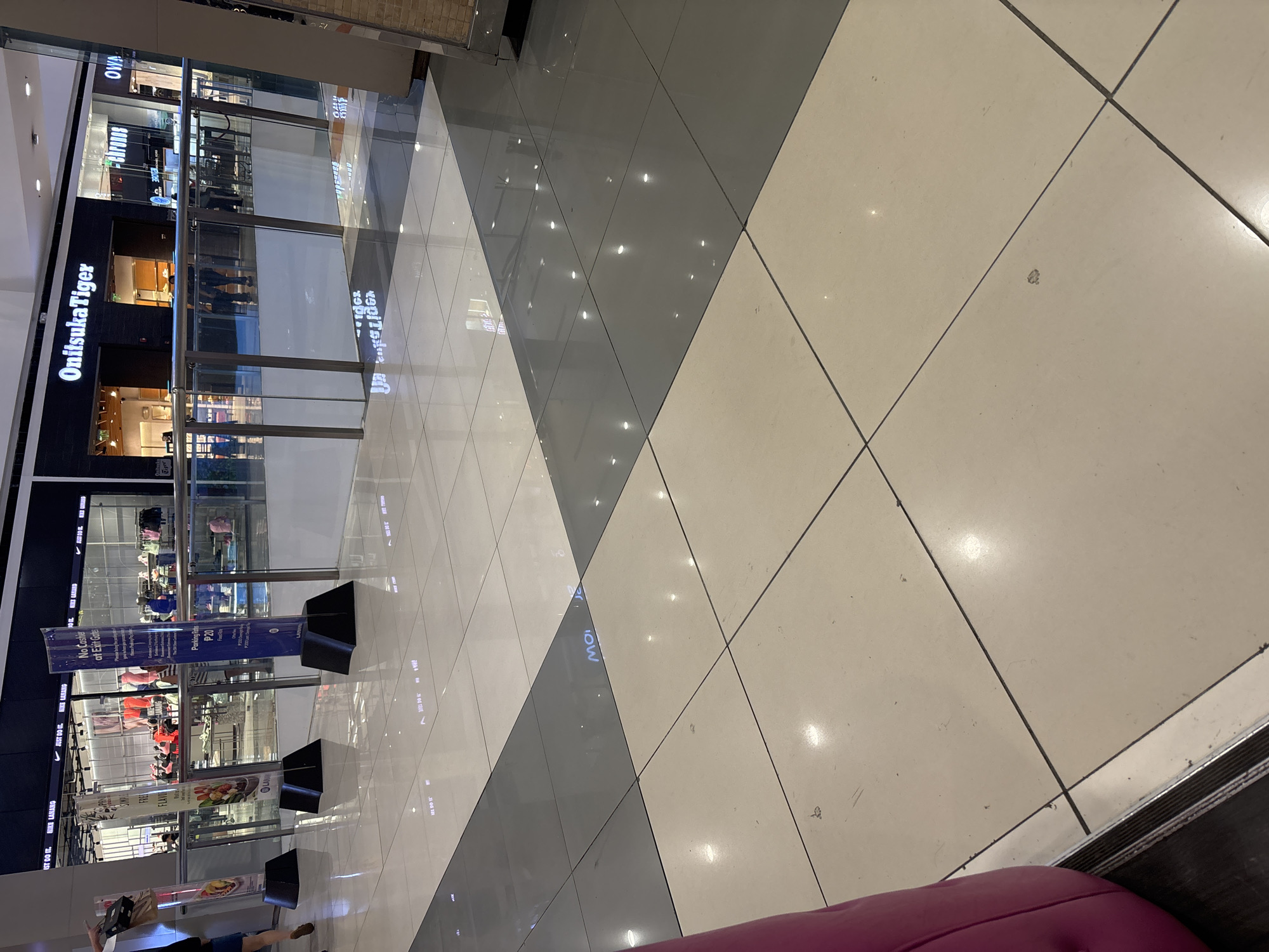 The image captures a view inside a brightly lit, modern commercial establishment, likely a shopping mall in Davao City, Philippines. The foreground displays a polished floor with alternating square tiles of light beige and dark gray, reflecting the overhead lights. Several vertical signs are visible, including one with blue and white text that reads "No Cashier at Exit Gates" and "Parking Rates P20 Fixed Rate." Another sign, a tri-fold banner, displays images of food items and text related to "Flavors." Behind these signs, a glass railing separates different levels of the establishment, beyond which are storefronts. One storefront prominently features the "Onitsuka Tiger" logo. Another displays the "Nike" logo. In the background, individuals can be seen browsing merchandise and walking, their presence adding a sense of activity to the scene. The overall impression is that of a clean, well-maintained commercial space.