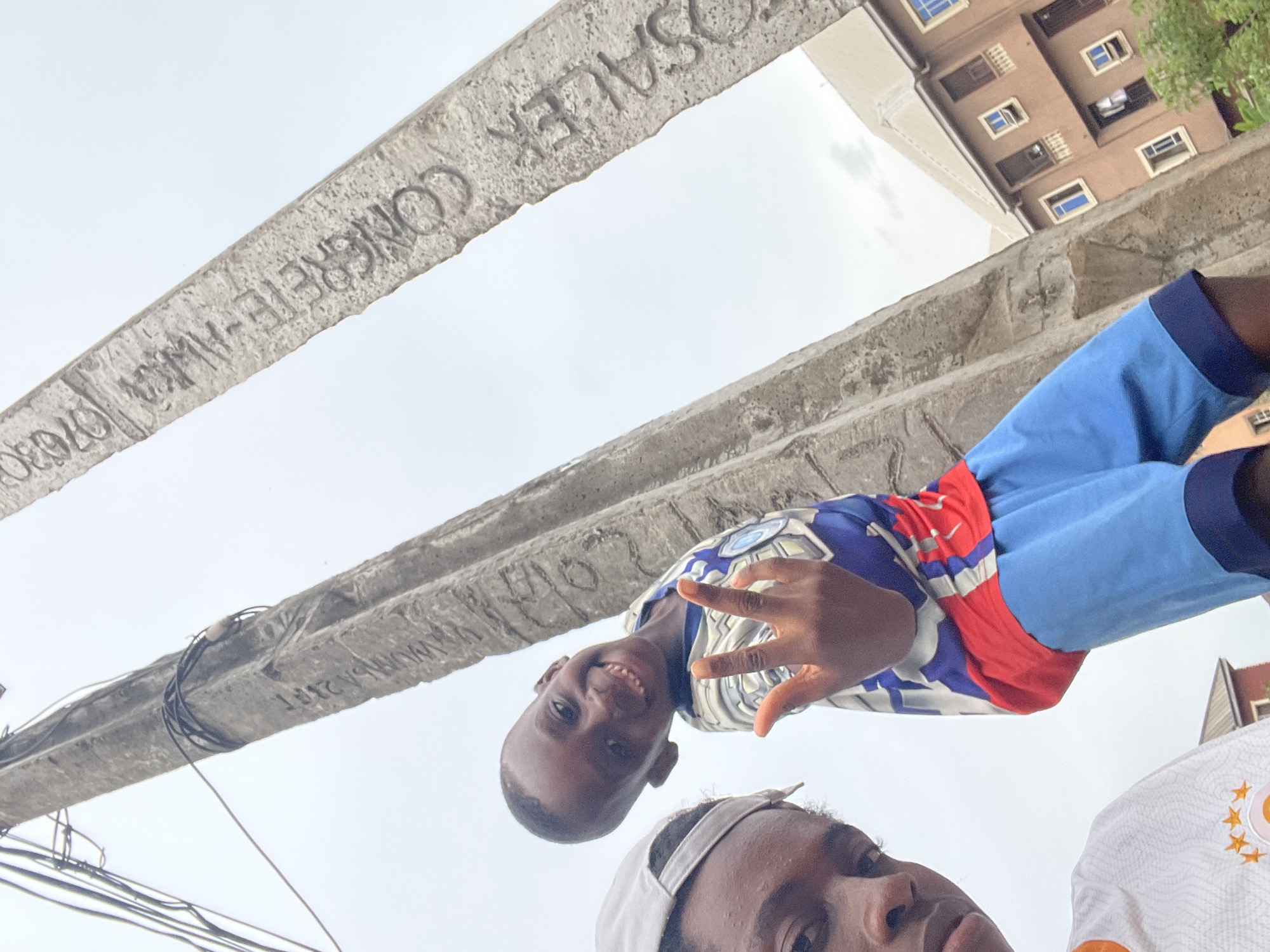 The image is an upward-facing shot of two people, likely a selfie, taken beneath two concrete beams in Onitsha, Nigeria. The writing on the concrete beams reads "CONCRETE-AWKA" and "ROSALEK CONCRETE". The taller building with multiple windows is visible in the background. The child is smiling and holding up four fingers. The other person is wearing a white baseball cap. A building is visible in the background. A web of black cables is attached to the lower concrete beam.