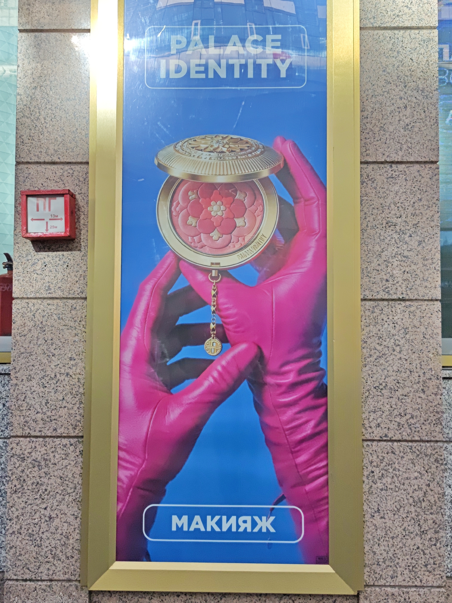 The image shows a large advertising poster on a building exterior in Moscow, Russia. The poster features a close-up of a person's hands wearing bright pink gloves, holding open a golden compact powder case with a floral design. The background of the poster is a vibrant blue. At the top of the poster, text reads "PALACE IDENTITY". At the bottom, in a white oval shape, is Russian text that reads "МАКИЯЖ", which translates to "MAKEUP". To the left of the poster, a red fire safety box is mounted on the stone wall of the building. The overall impression is of a cosmetic advertisement.