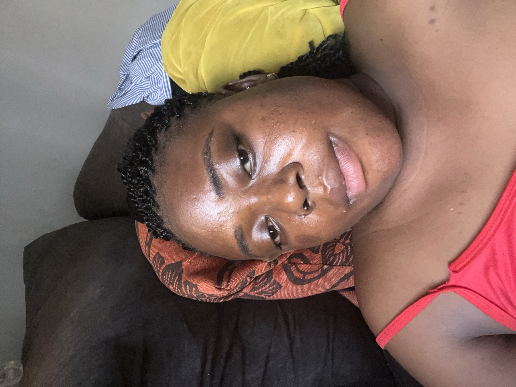 The media file is a close-up, slightly over-the-shoulder shot of a young Black woman lying down. She is positioned on her side, with her head resting on a patterned pillow and a dark, textured surface, possibly a sofa or bed. Behind her, a yellow fabric is visible, and further back, a striped blue and white fabric is also present, suggesting clothing or bedding.
The woman has braided hair and is wearing a red garment, possibly a top or swimwear, with thin straps. Her skin appears dewy, and she has a nose ring. She is looking towards the camera with a gentle smile and slightly parted lips, conveying a relaxed or content emotion. The lighting suggests it might be daytime, possibly indoors, as there are no strong shadows or indications of outdoor weather. The scene is intimate and personal, a candid portrait. There is no visible text in the image. The location context provided is Abuja, Nigeria.