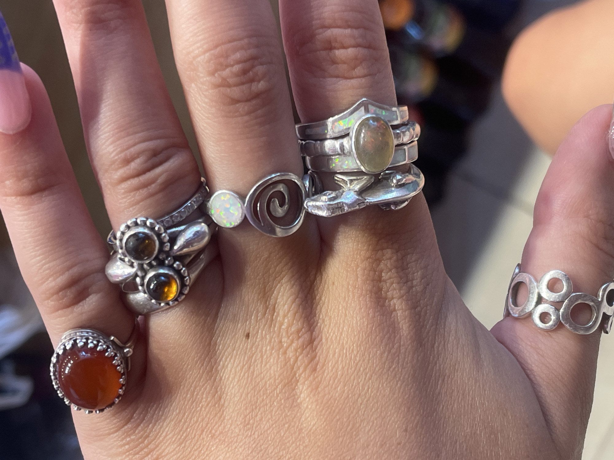 The image shows a close-up view of a person's hand adorned with multiple rings. The rings are made of silver and feature various gemstones, including a prominent reddish-orange stone on the thumb, two amber-colored stones on a ring resembling an owl, and opal stones on rings stacked on the middle finger. One ring on the middle finger is a stack of thin silver bands, with an oval opal stone on the top band and smaller opals integrated into the sides of the bands. Another ring on the middle finger is a silver spiral design. A ring on the index finger has a silver lizard or gecko design. On the pinky finger, there is a silver ring with a geometric pattern of interconnected circles of varying sizes. The background is blurred, but appears to be an indoor setting with hints of colored lights, possibly from a shop or display. There is no identifiable information within the image that places it in Raanana, Israel, or any specific city.